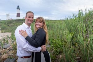 Smiling couple posing together near a red and white lighthouse surrounded by tall green grass.