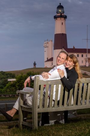 Happy couple sitting on a wooden bench laughing with a tall lighthouse behind them at dusk.