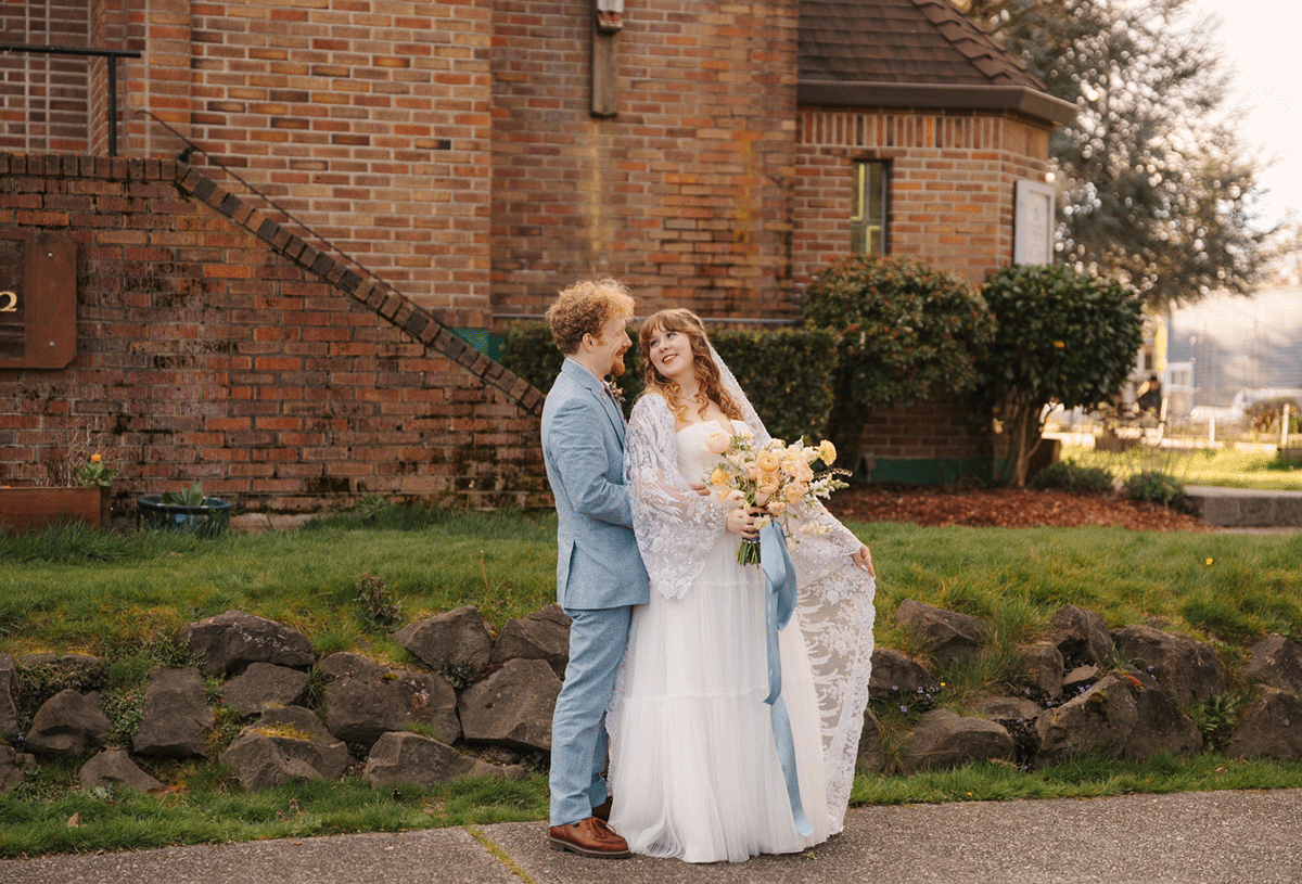 Micah and Sage embrace outdoors near a brick church, she in a white gown, he in a blue suit for their Capitol Hill Seattle Wedding.