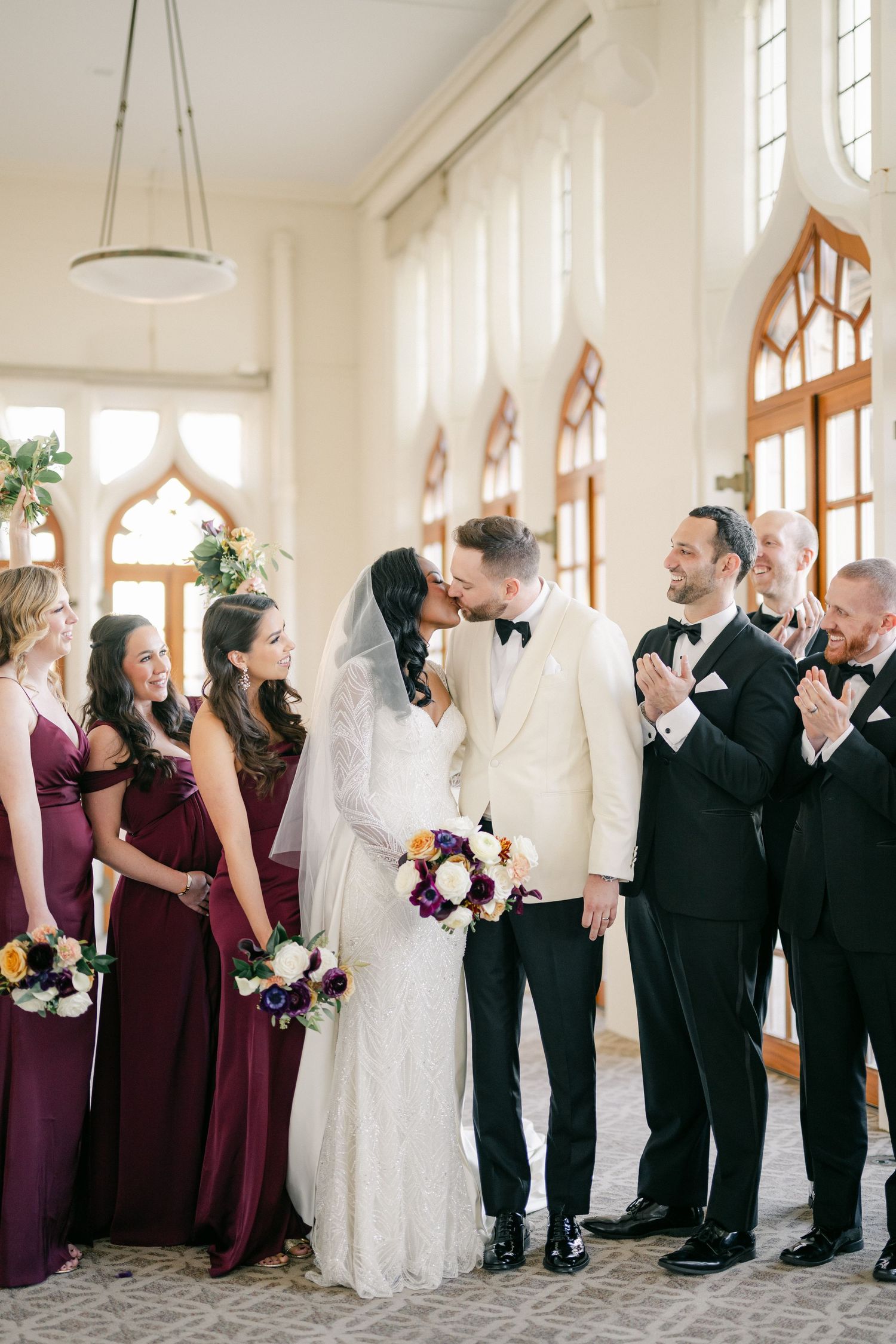 Bride and groom share first kiss surrounded by bridesmaids in burgundy gowns. - University Club of Chicago Wedding