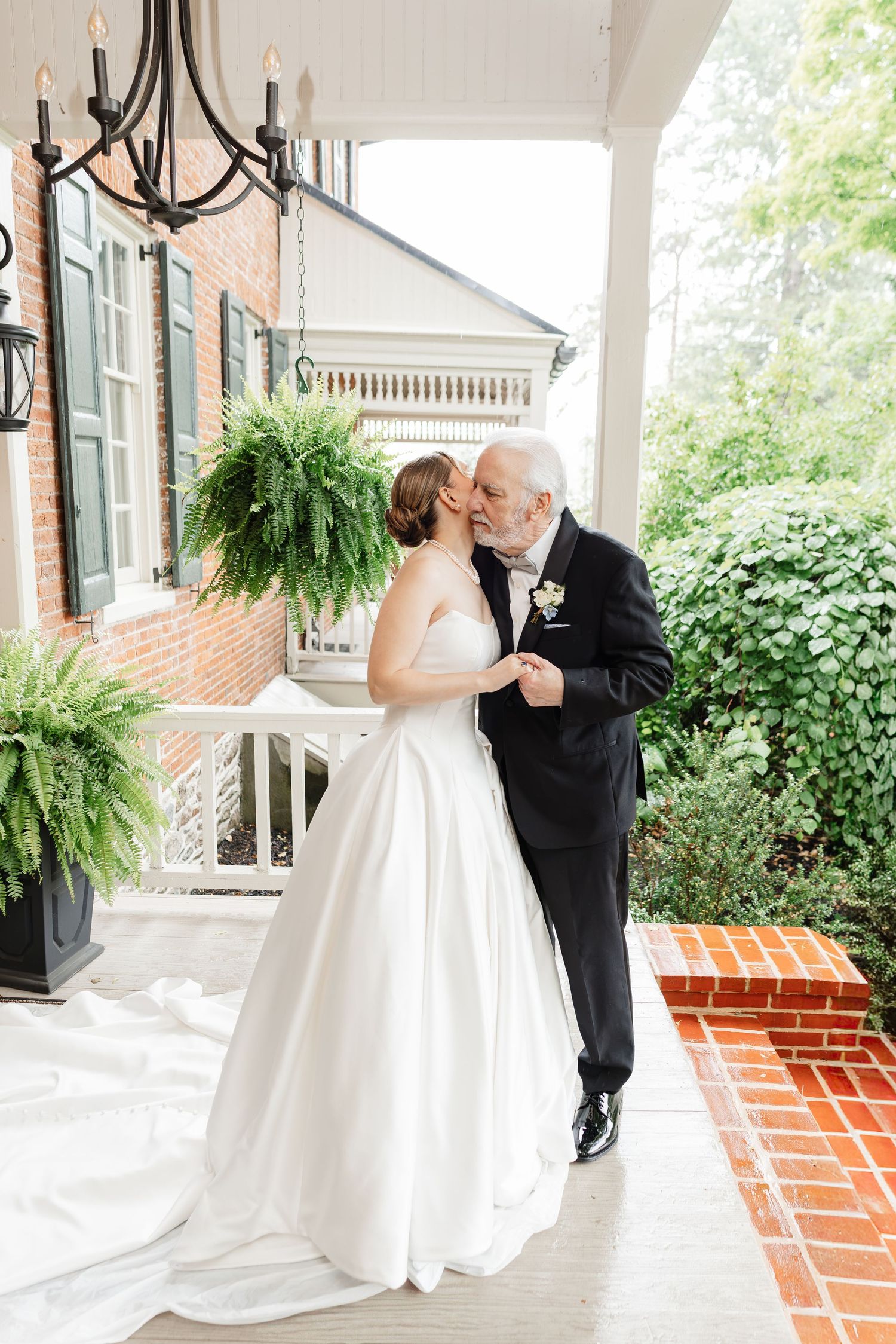 Bride and older gentleman share a tender kiss on a brick porch of a historic home surrounded by lush ferns.