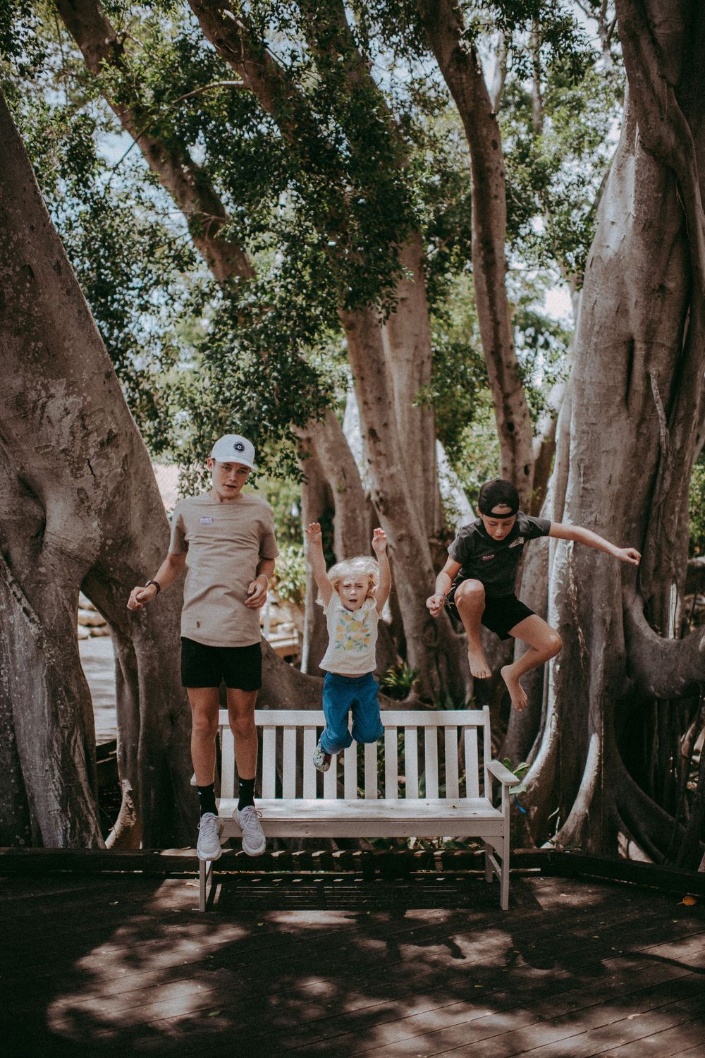Family of three joyfully jumping off a white bench surrounded by large trees in a sunny park.