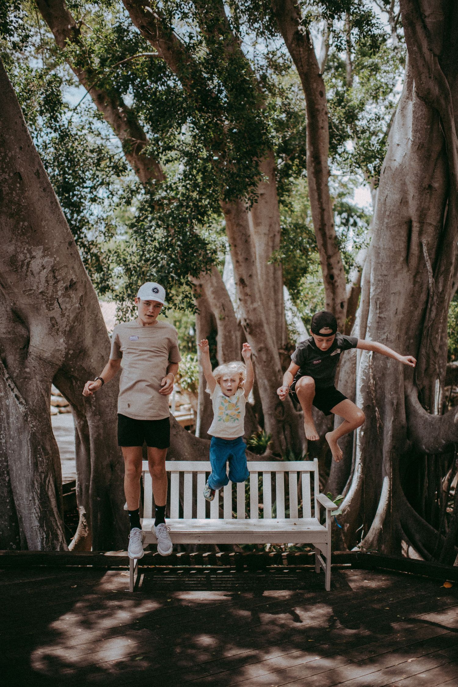 Family of three joyfully jumping off a white bench surrounded by large trees in a sunny park.
