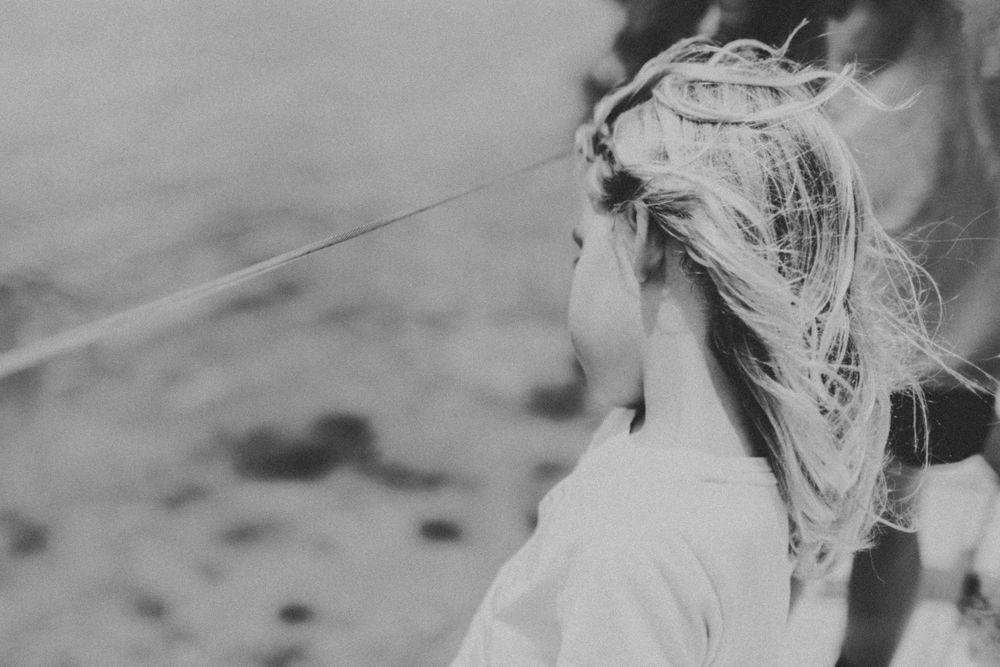 Black and white photo of a blonde girl with windswept hair outdoors near a beach.
