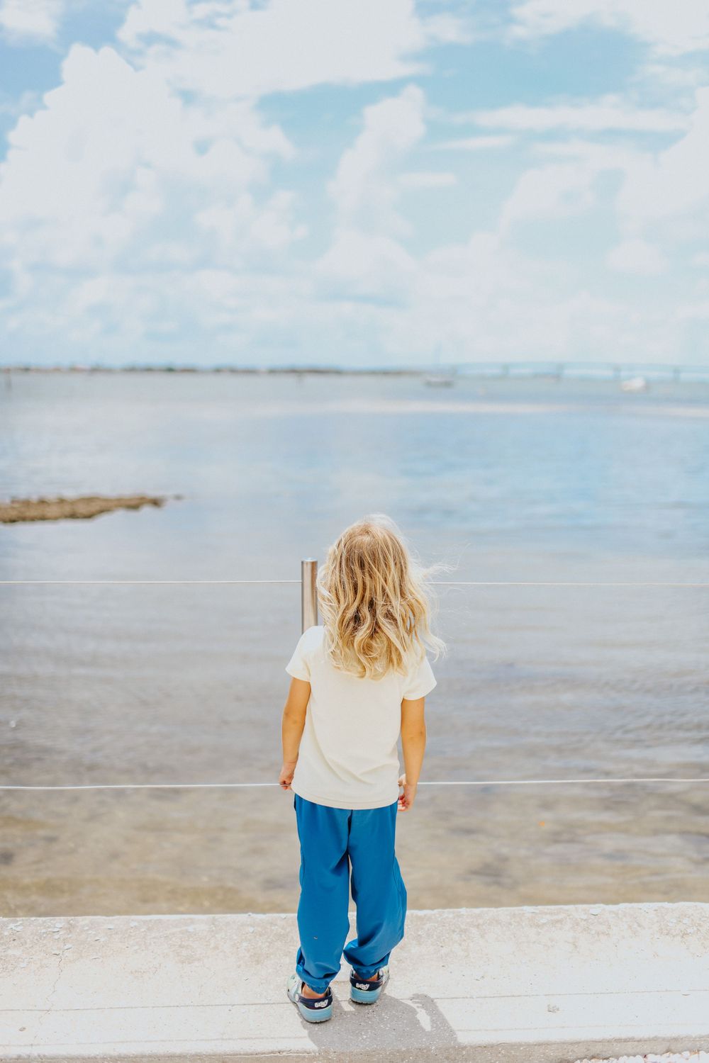 Young blonde child in white tee and blue pants stands on waterfront ledge gazing at calm ocean under cloudy sky.