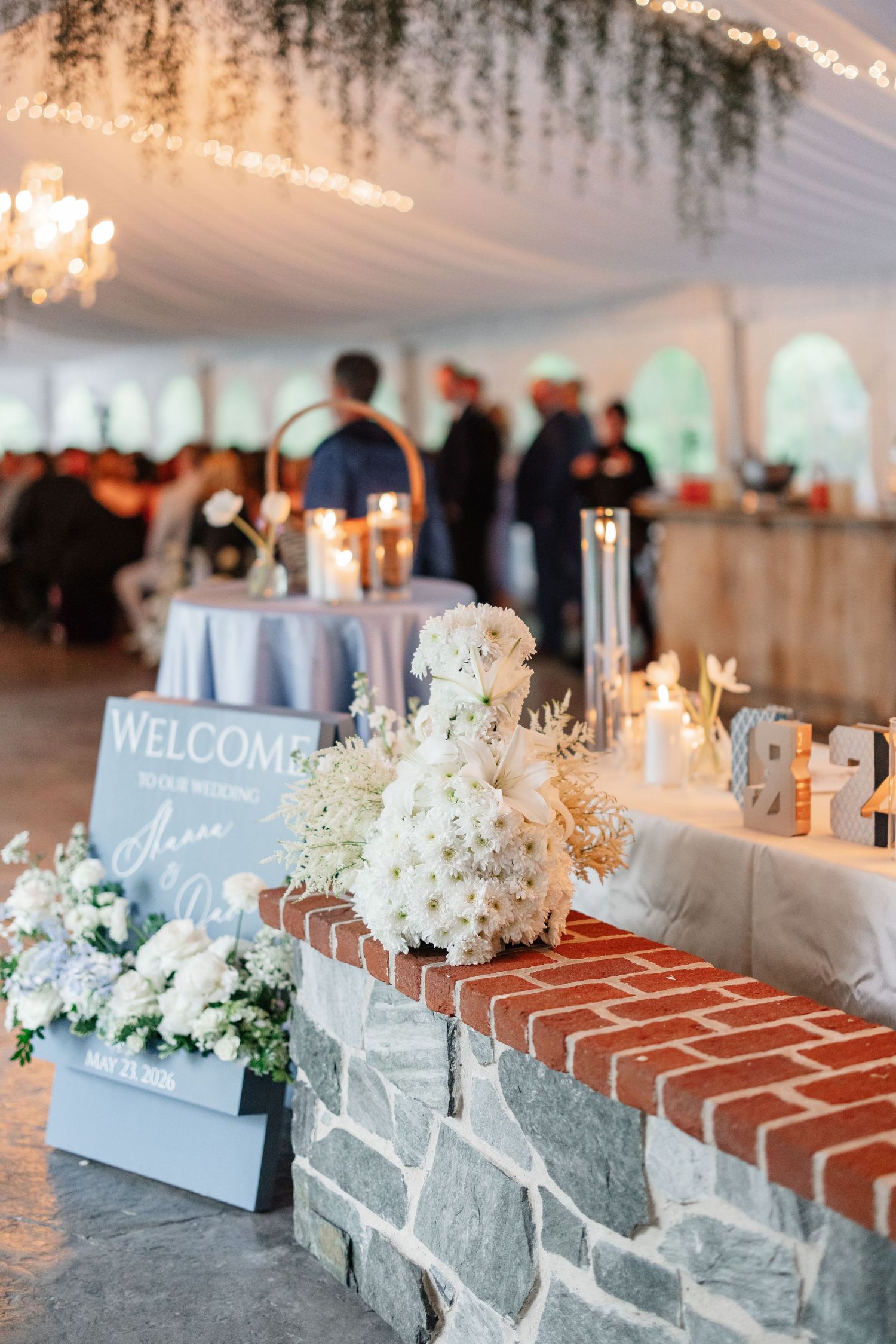 Wedding welcome sign with white floral arrangements on stone ledge inside elegant tented venue.