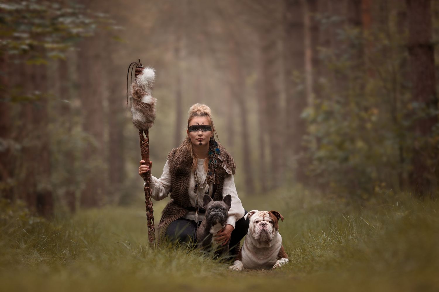 Woman in warrior costume holding a staff with feathers, sitting with two bulldogs in a misty forest.