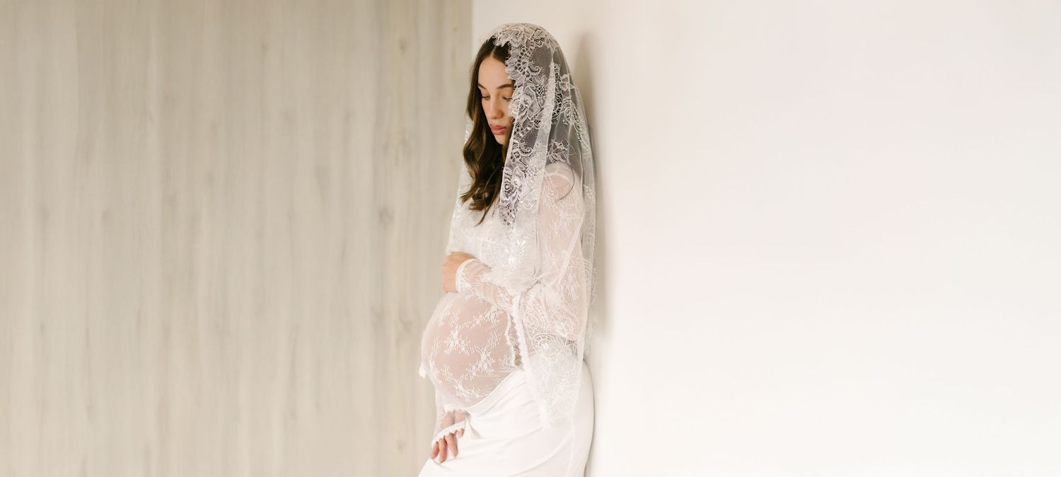 Expectant mother in a lace gown and long veil beside a bright studio wall.