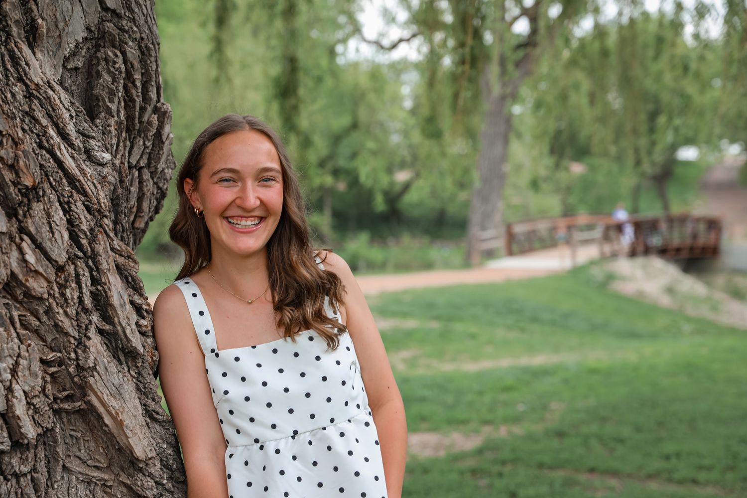 Young woman in floral dress smiling by a peaceful creek surrounded by lush green trees.