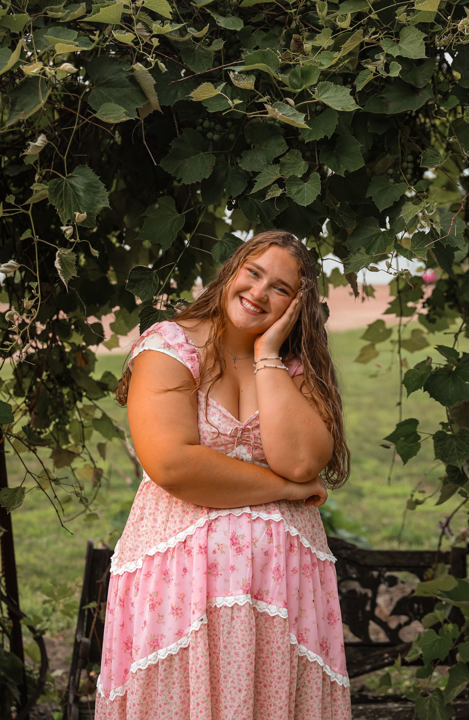 Smiling young woman in pink floral dress posing under green leafy arbor in outdoor garden setting.