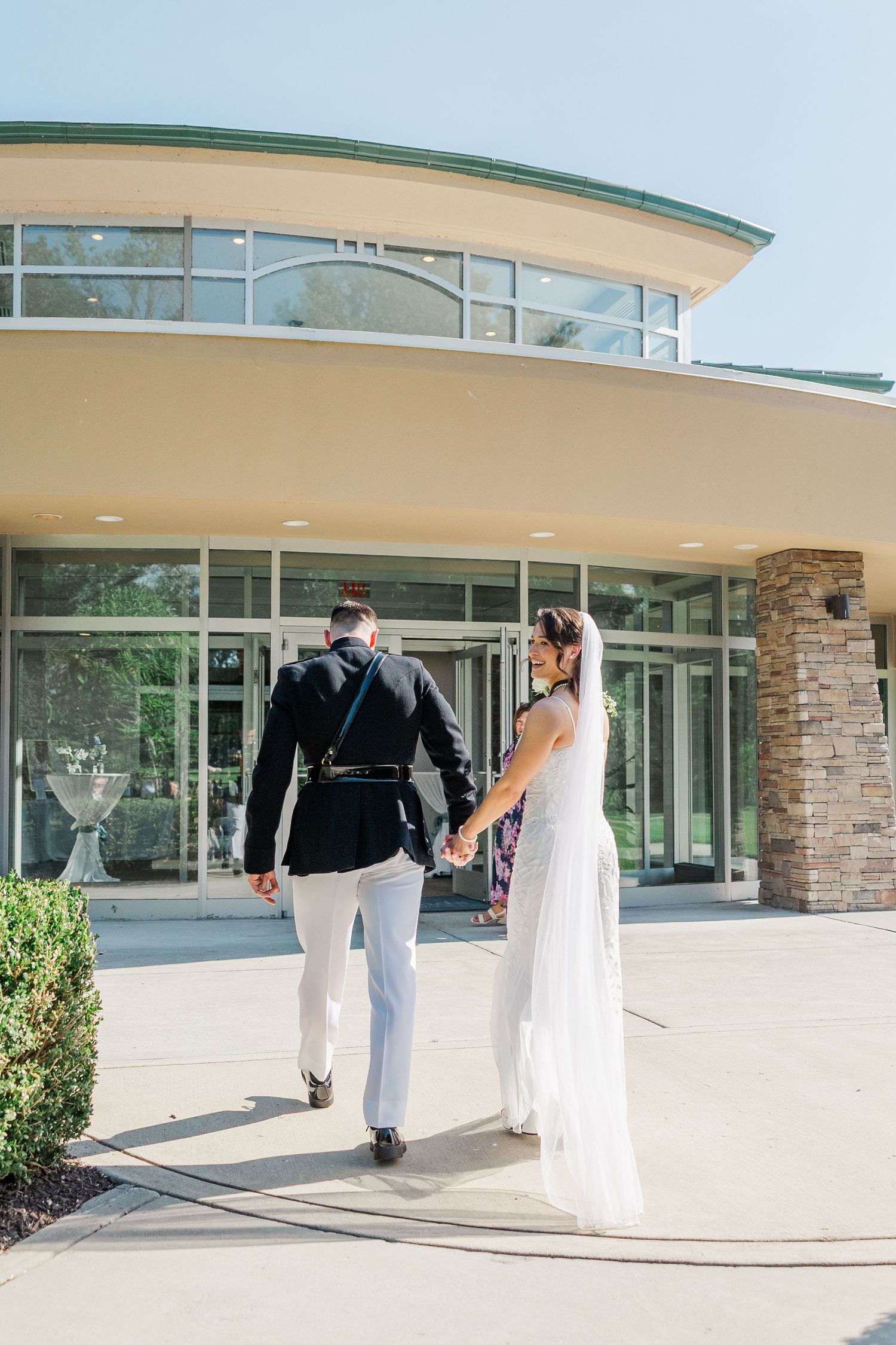 A bride and groom walk hand-in-hand toward an elegant venue entrance on their sunny wedding day.