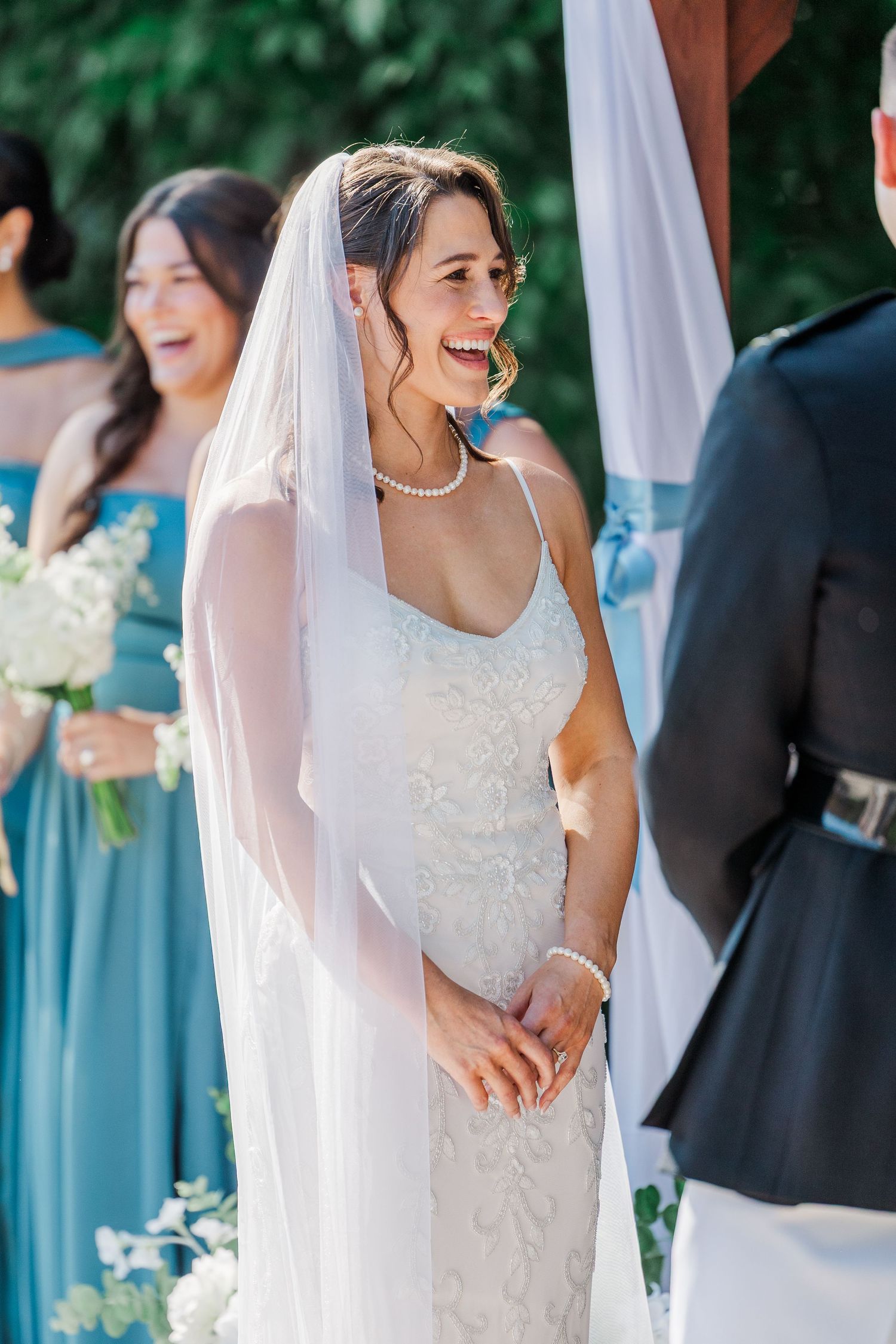 Bride in white lace gown and veil laughing joyfully at outdoor wedding ceremony with bridesmaids in blue.