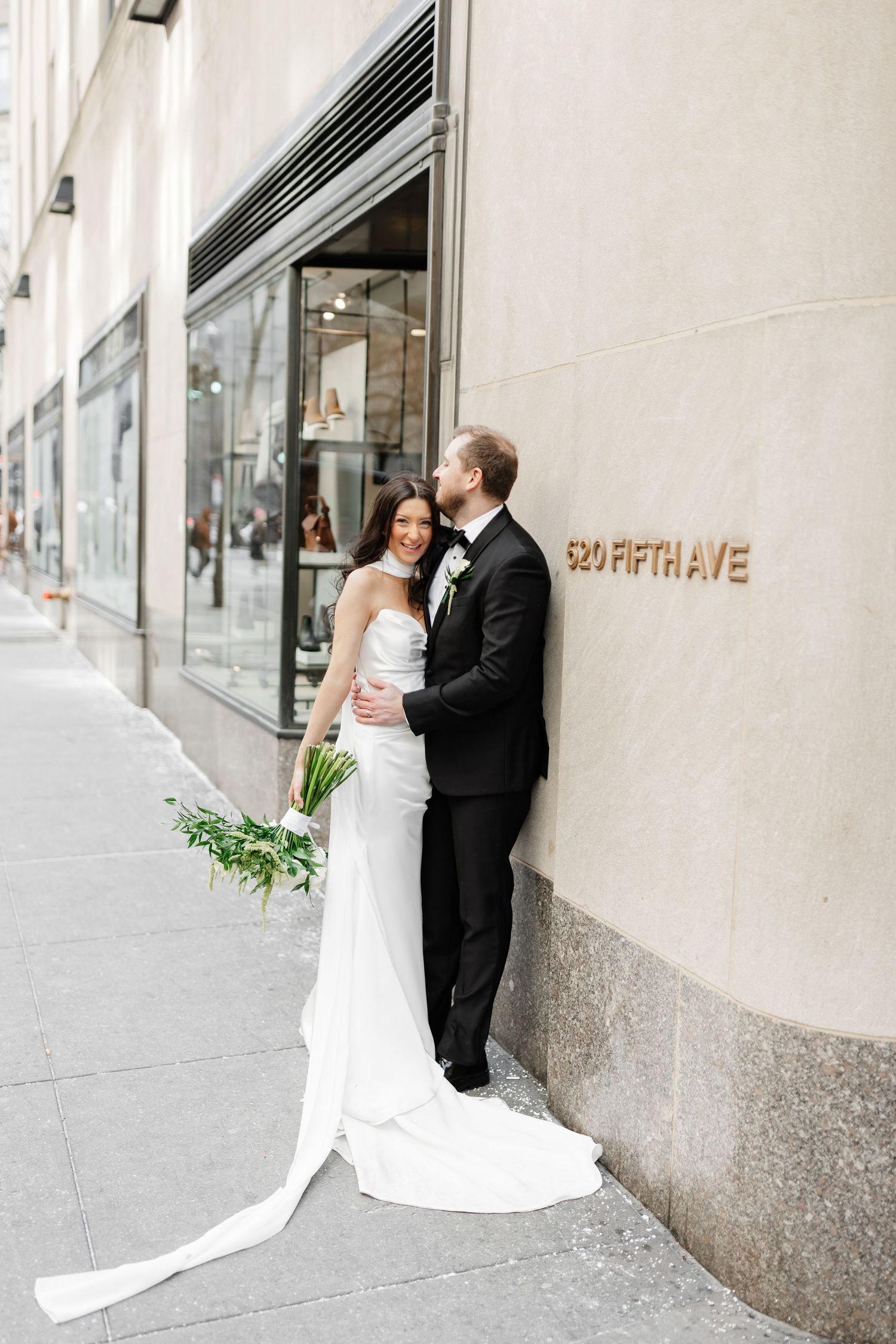 Bride and groom pose against 520 Fifth Ave building in NYC, bride in white gown holding greenery bouquet.