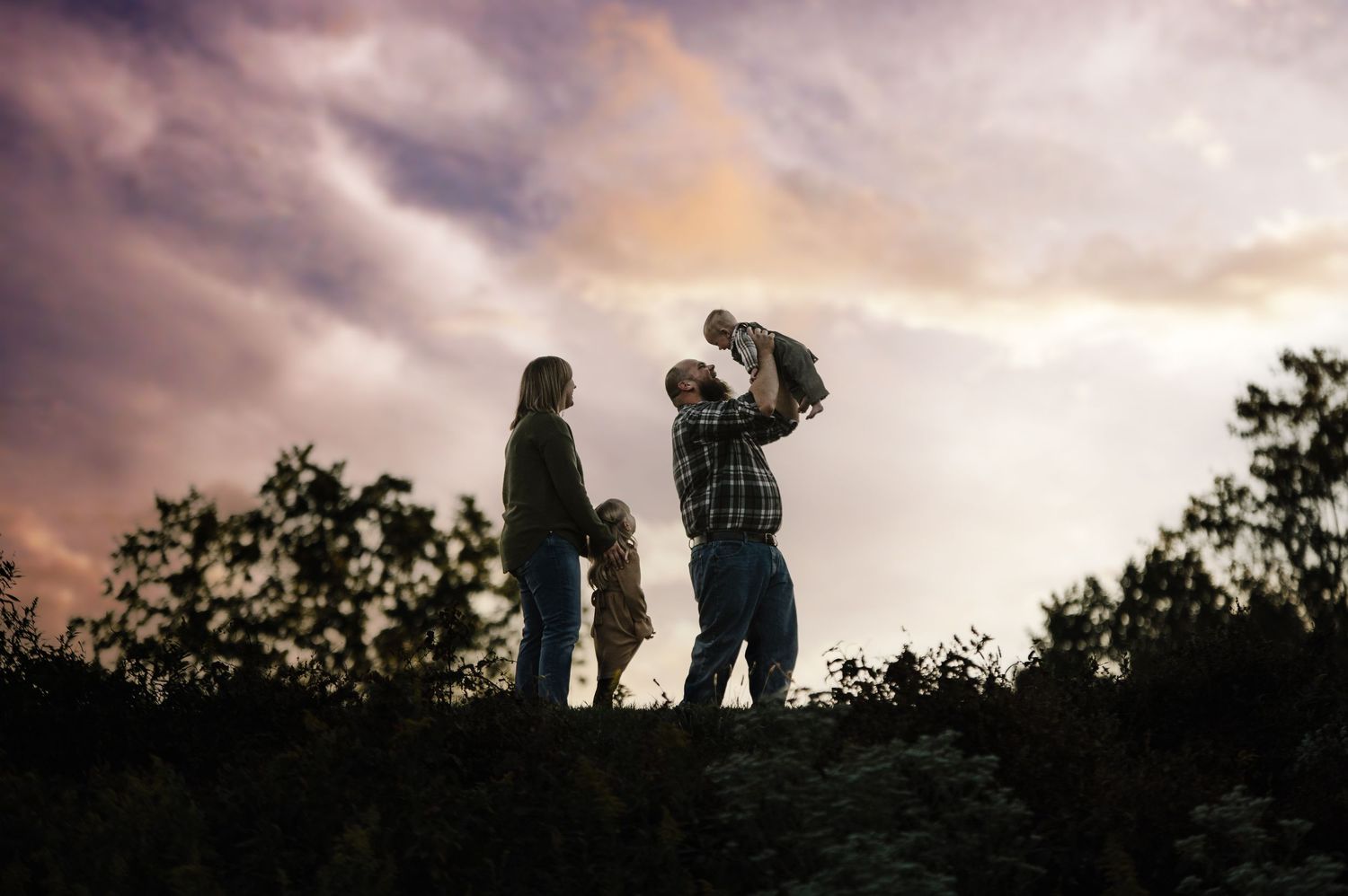 Silhouette of a family at sunset, parents with two children, one lifted high, against a dramatic cloudy sky.