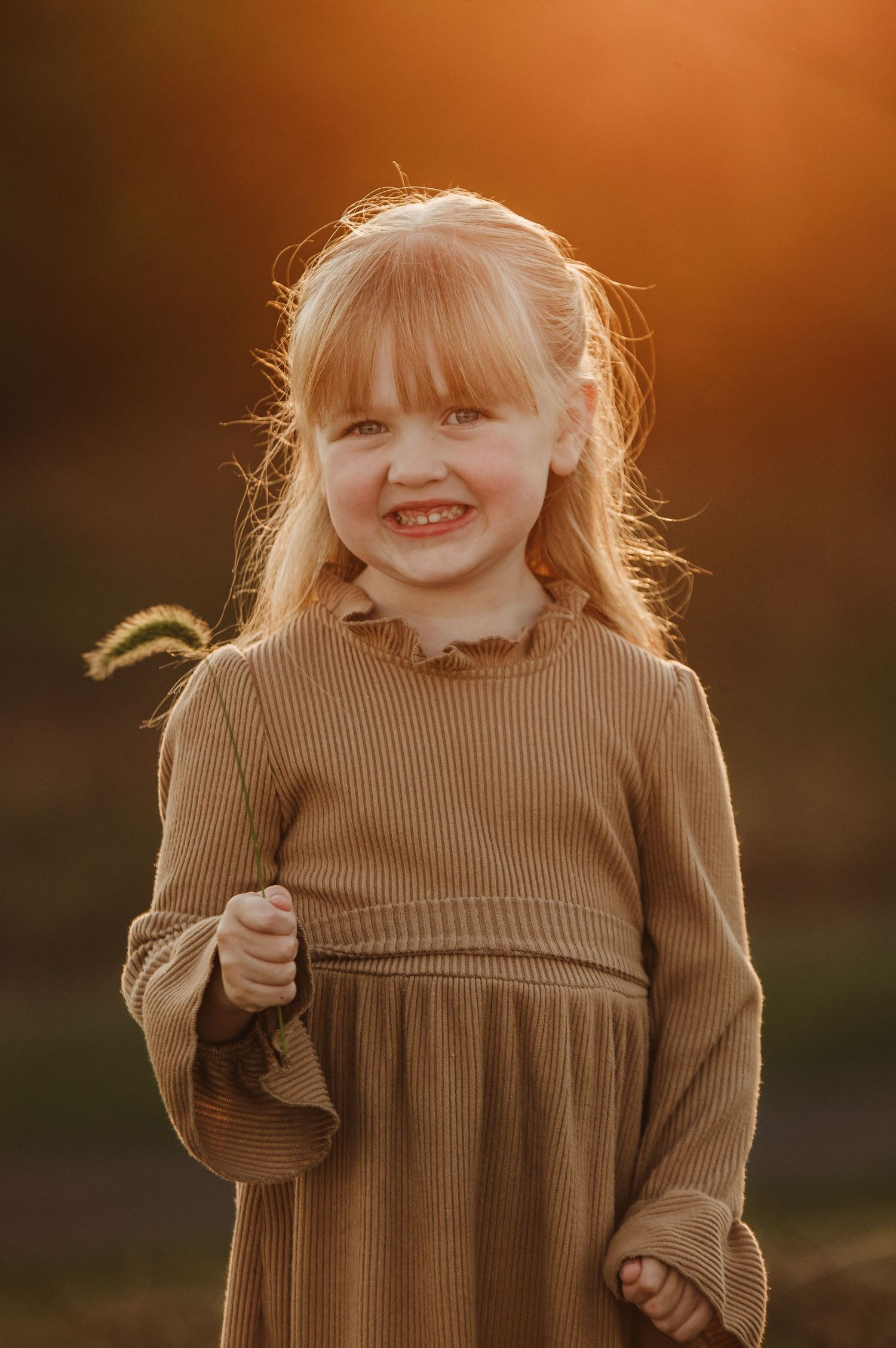 Smiling blonde toddler girl in brown ribbed dress outdoors in golden sunset light.