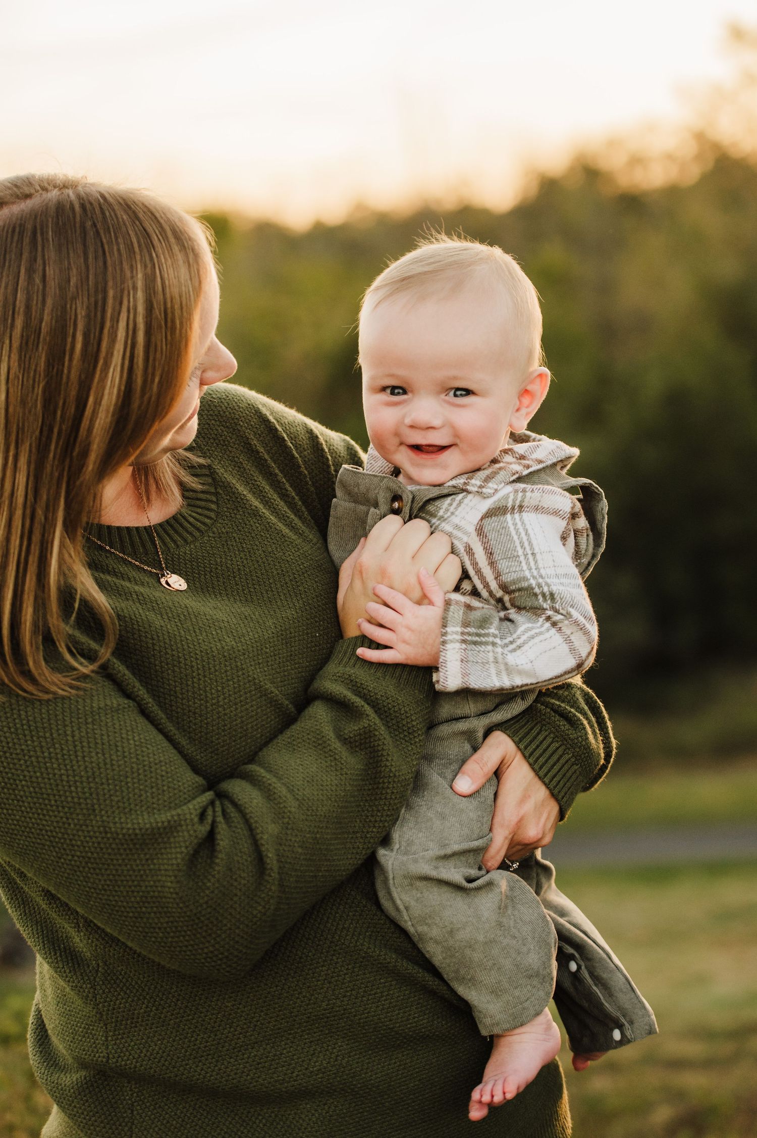 A mother in a green sweater holds her smiling baby boy outdoors at golden hour.