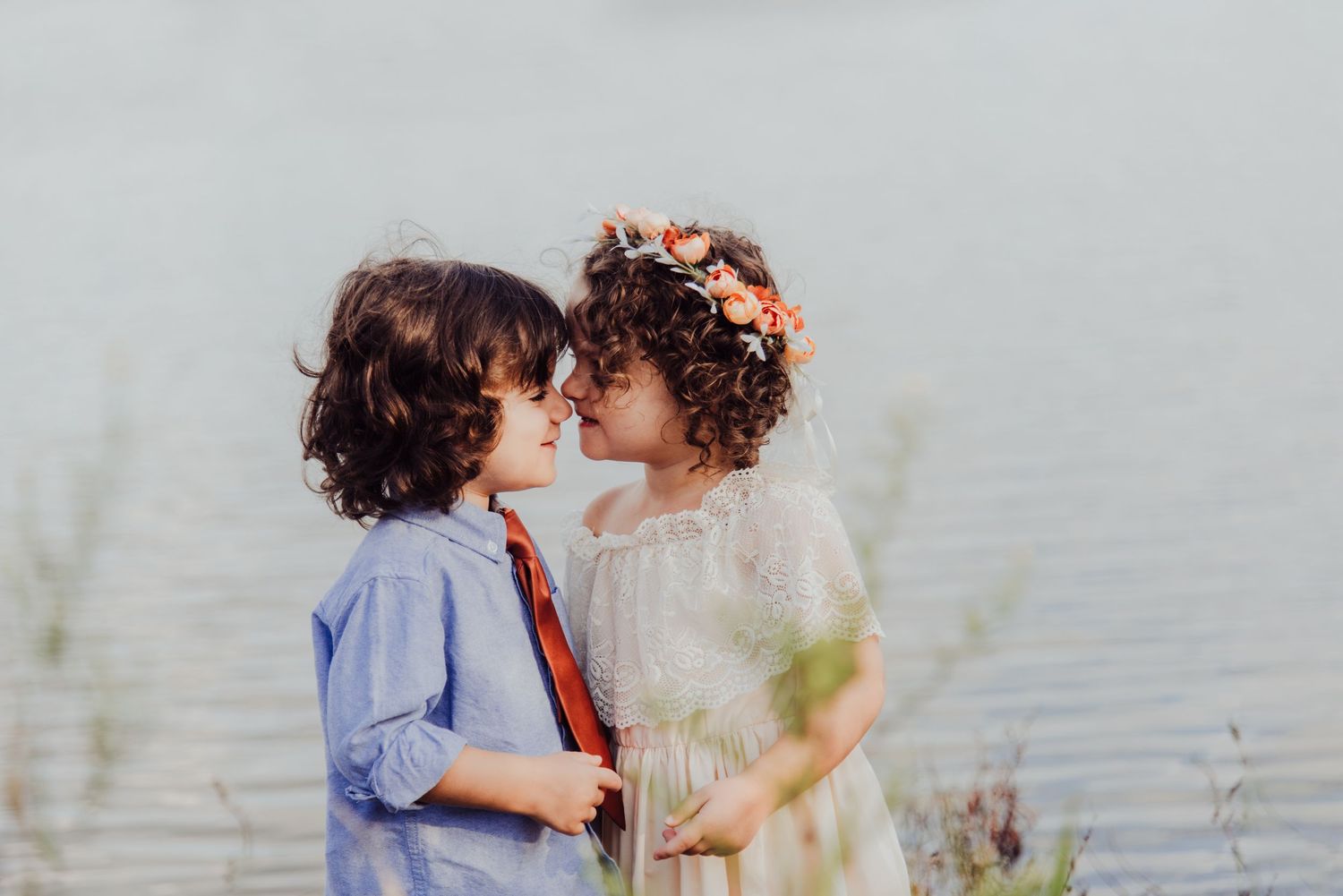 Two young children in elegant attire share a sweet moment near a serene lake, with a girl wearing a floral crown.