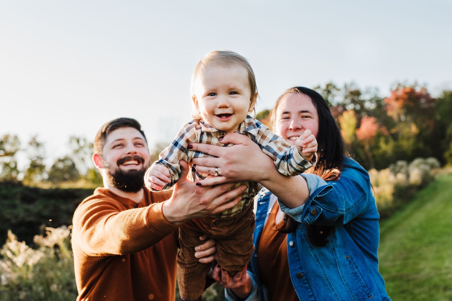 Happy family with parents holding smiling baby outdoors in a sunny autumn park setting.