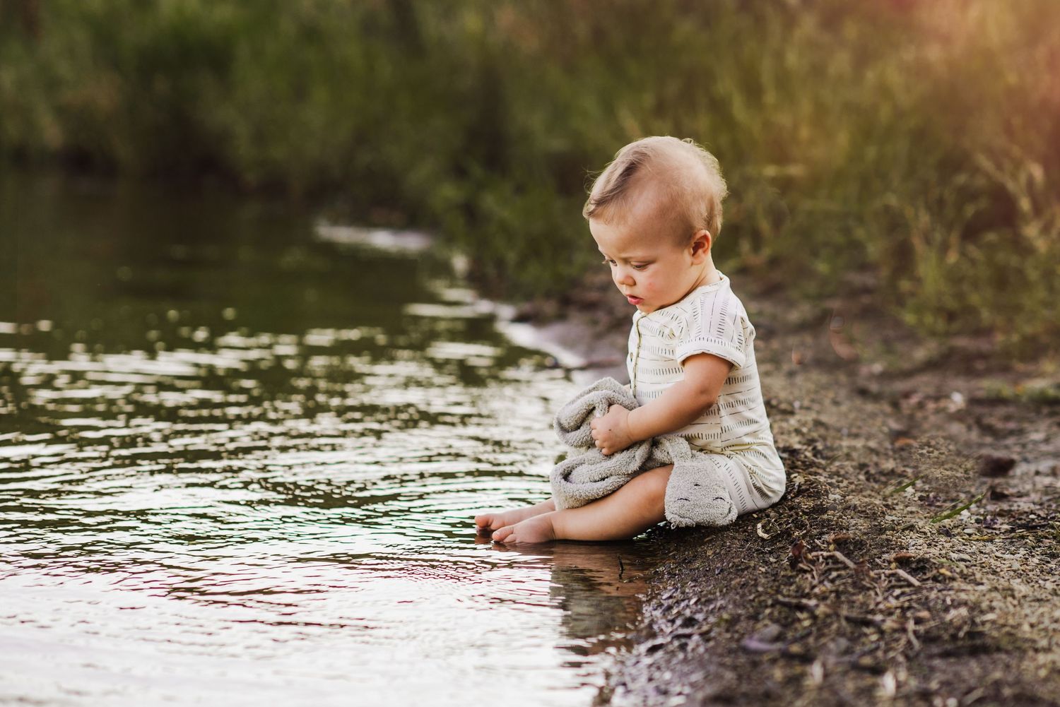 Toddler sitting by water's edge touching the surface, wearing a light romper in a serene outdoor nature setting.