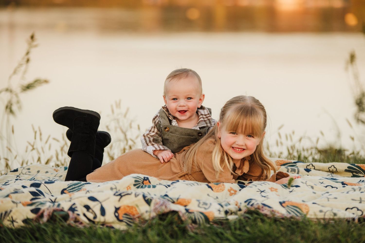 Two young siblings smiling on a floral blanket by a lake at sunset.