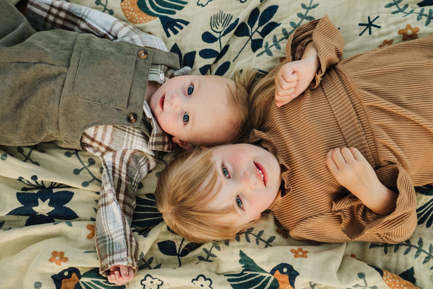 Two young children lying on a floral patterned blanket, wearing cozy earth-toned outfits, looking up at the camera.