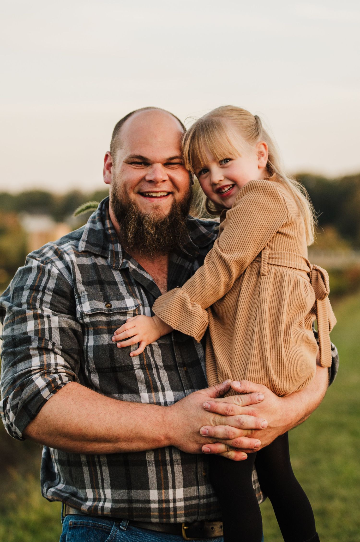 A bearded man in a plaid shirt smiles while holding a young blonde girl outdoors in a sunny field.