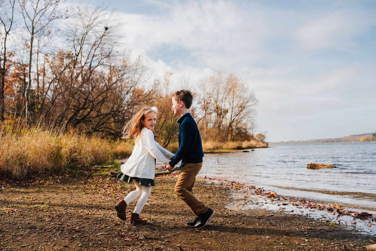Couple holding hands and dancing on an autumn lakeshore with colorful fall foliage in the background.