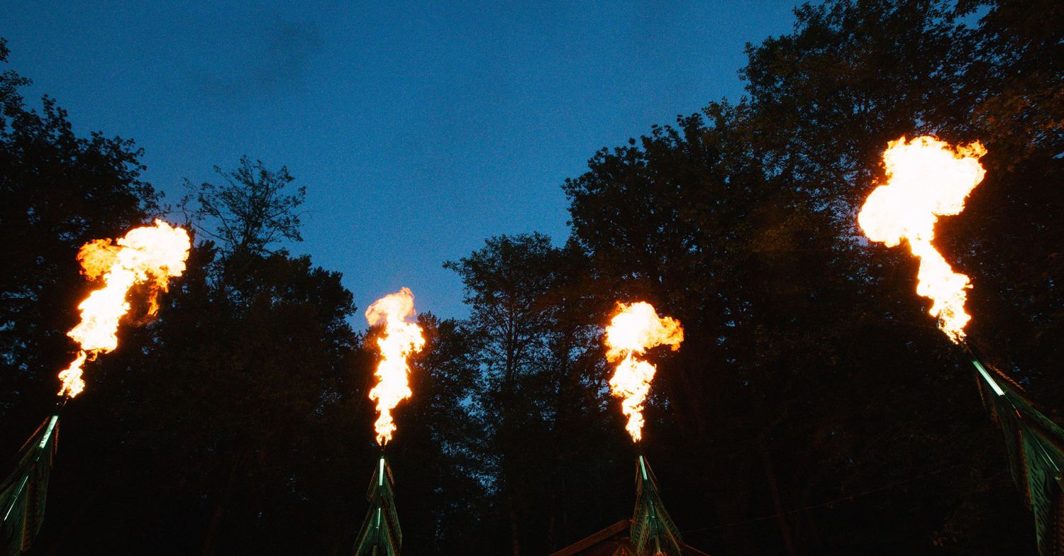 Multiple large flame torches blazing against a twilight sky with dark trees silhouetted in the background.