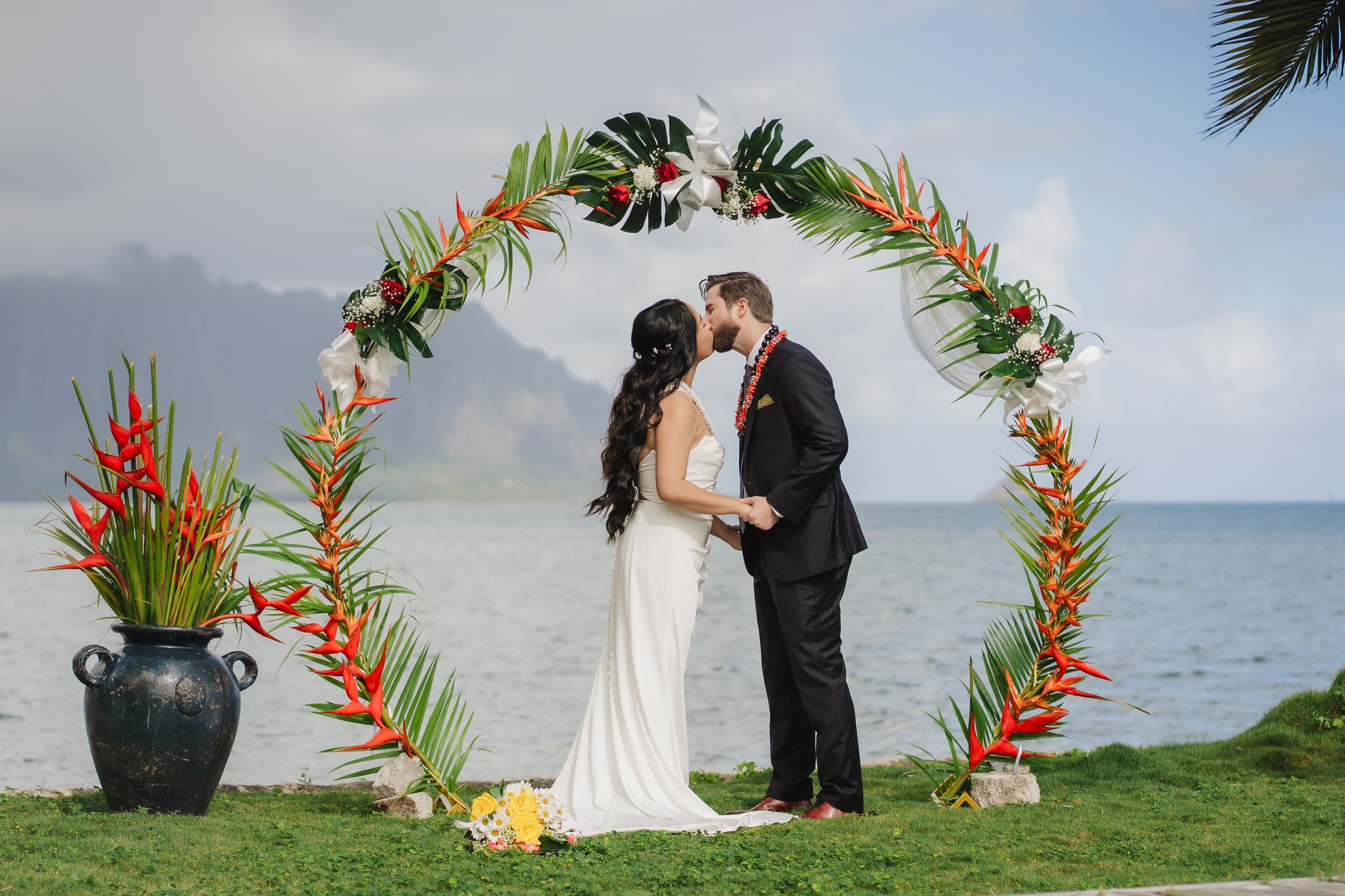 Couple kissing under tropical floral arch at beachside wedding ceremony in Hawaii with ocean and mountains backdrop.