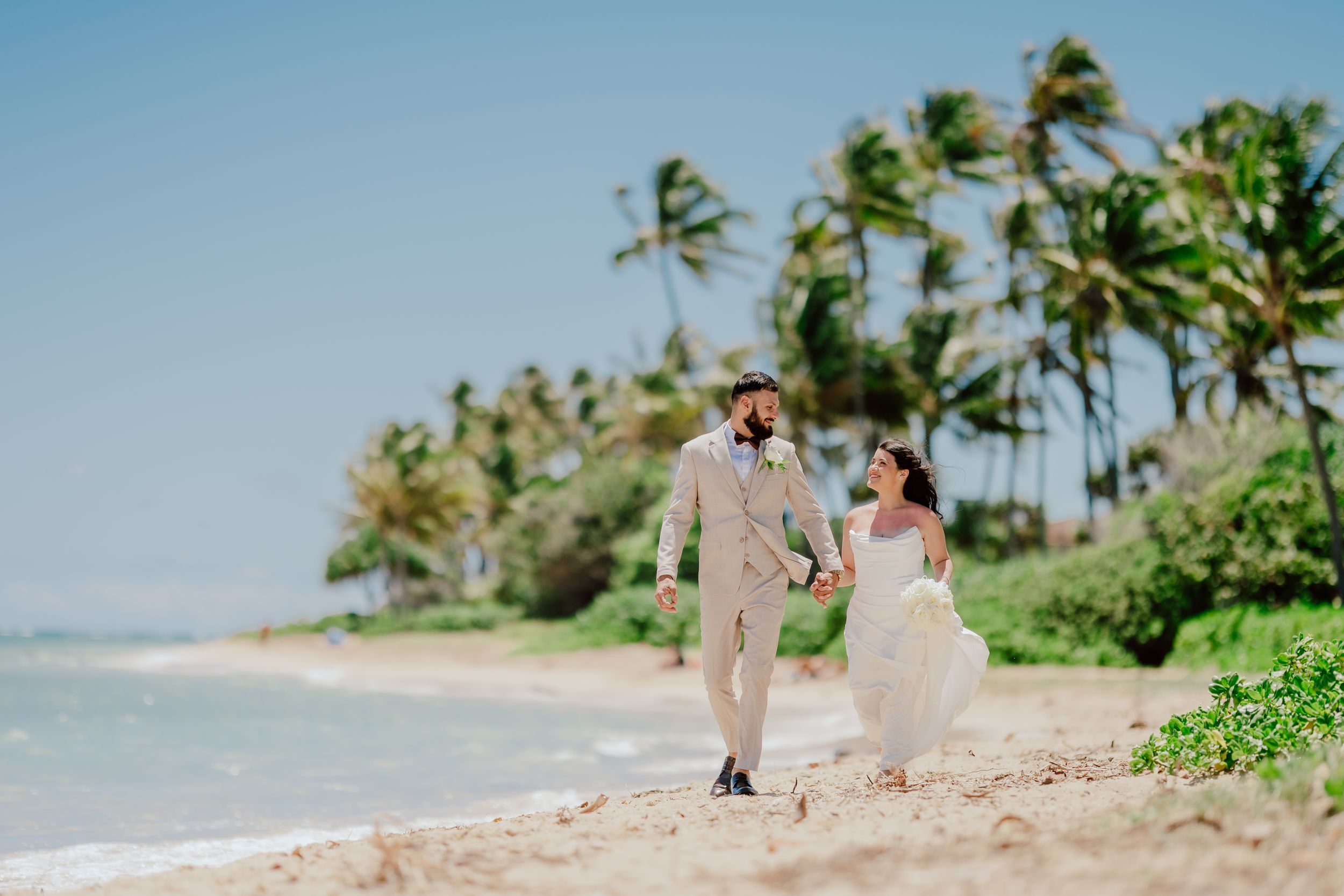 Bride and groom walking hand in hand on a tropical beach with palm trees swaying in the wind on their wedding day.