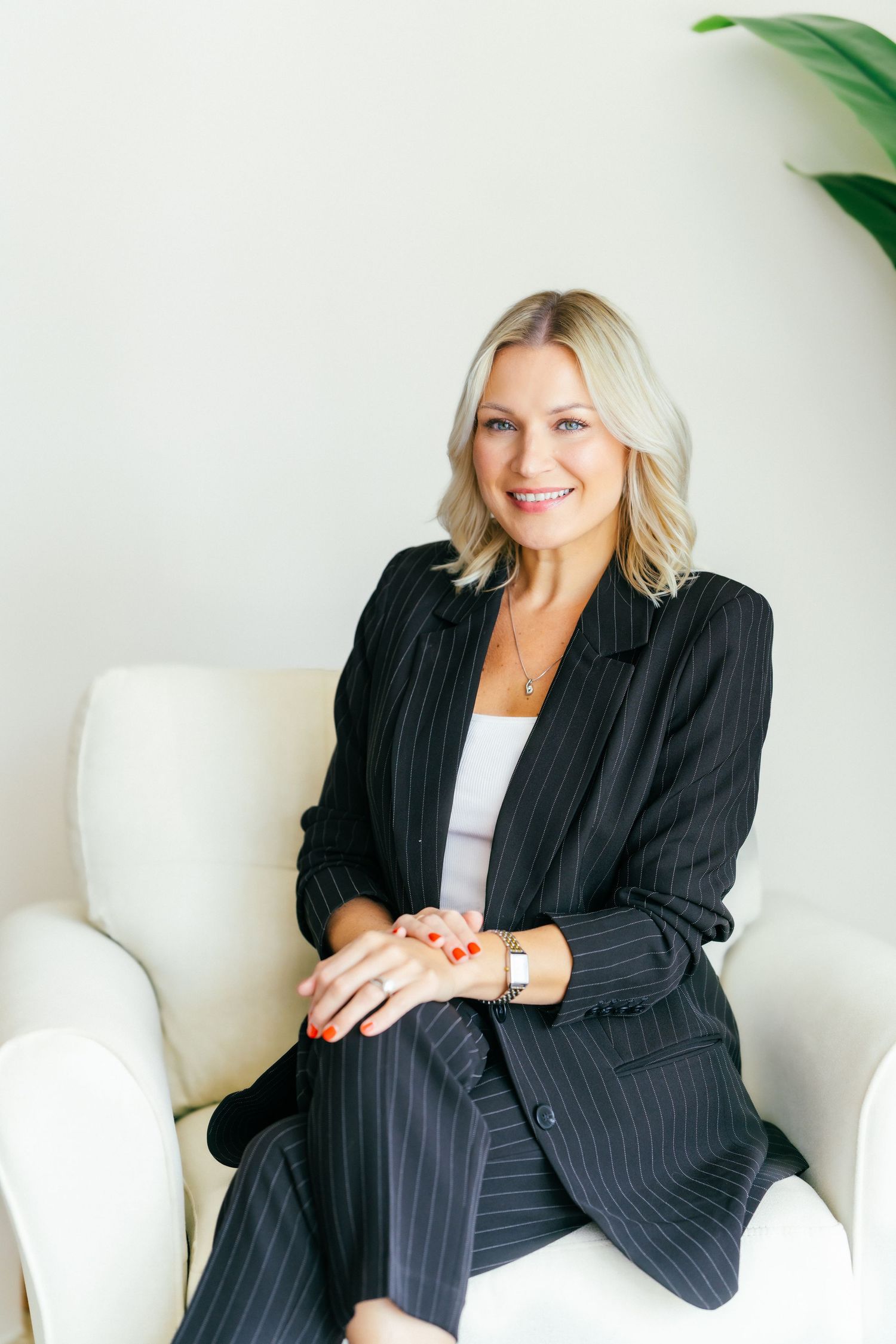 A smiling brunette woman in a beige blazer and shorts sits on a chair holding a tablet, wearing black heels.