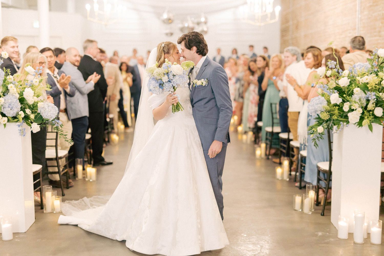 Bride and groom share a kiss in the aisle after their wedding ceremony, surrounded by guests and floral arrangements.