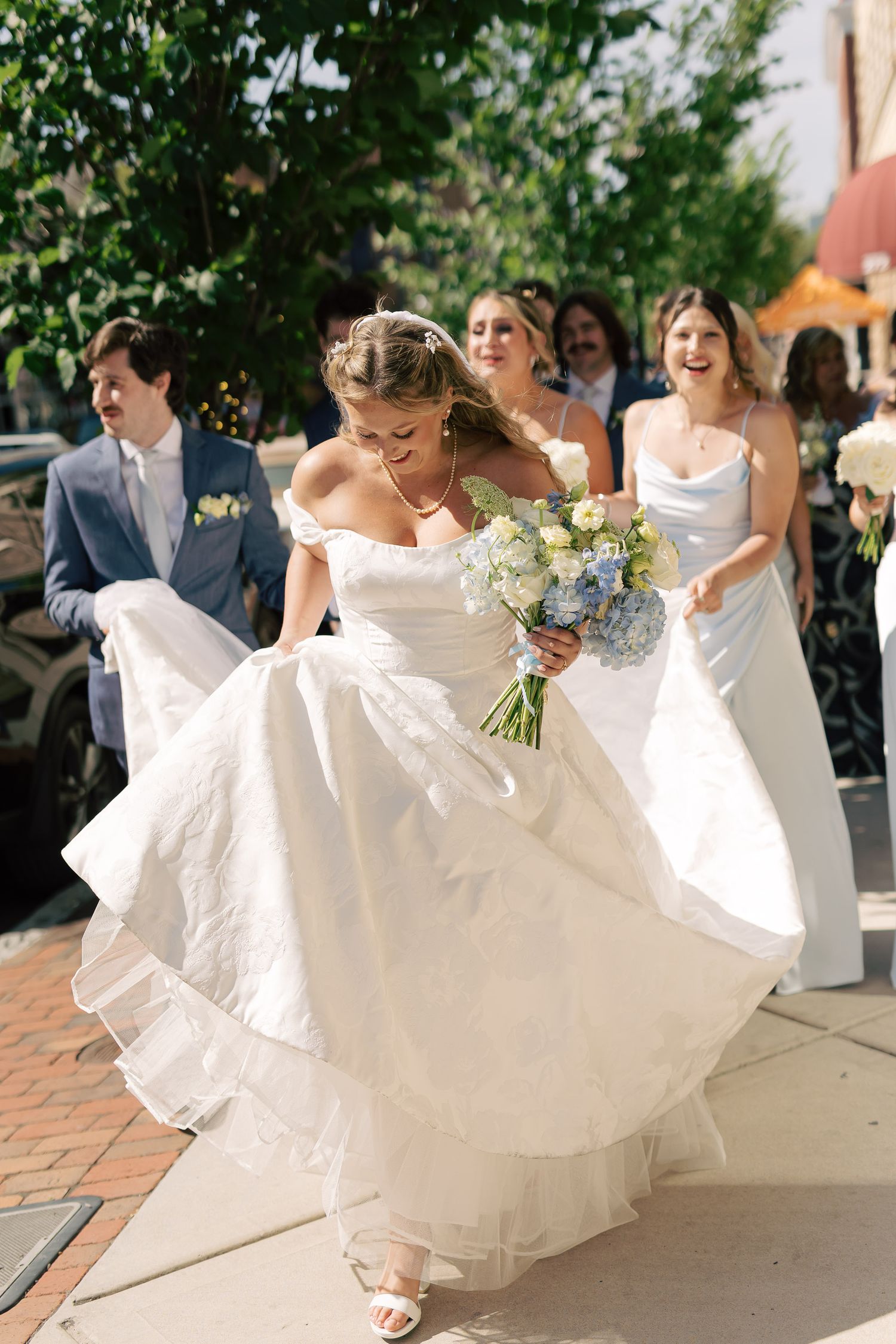 Radiant bride lifting her white ball gown while walking outdoors, holding a blue and white bouquet.