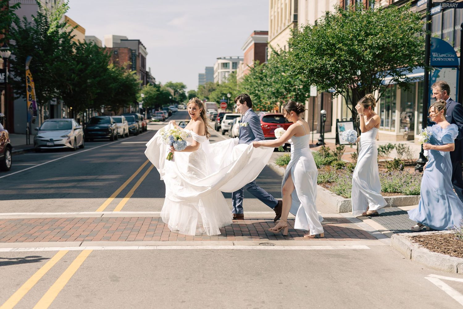Wedding party helps bride cross urban street, bridesmaids in blue dresses carrying white gown train.