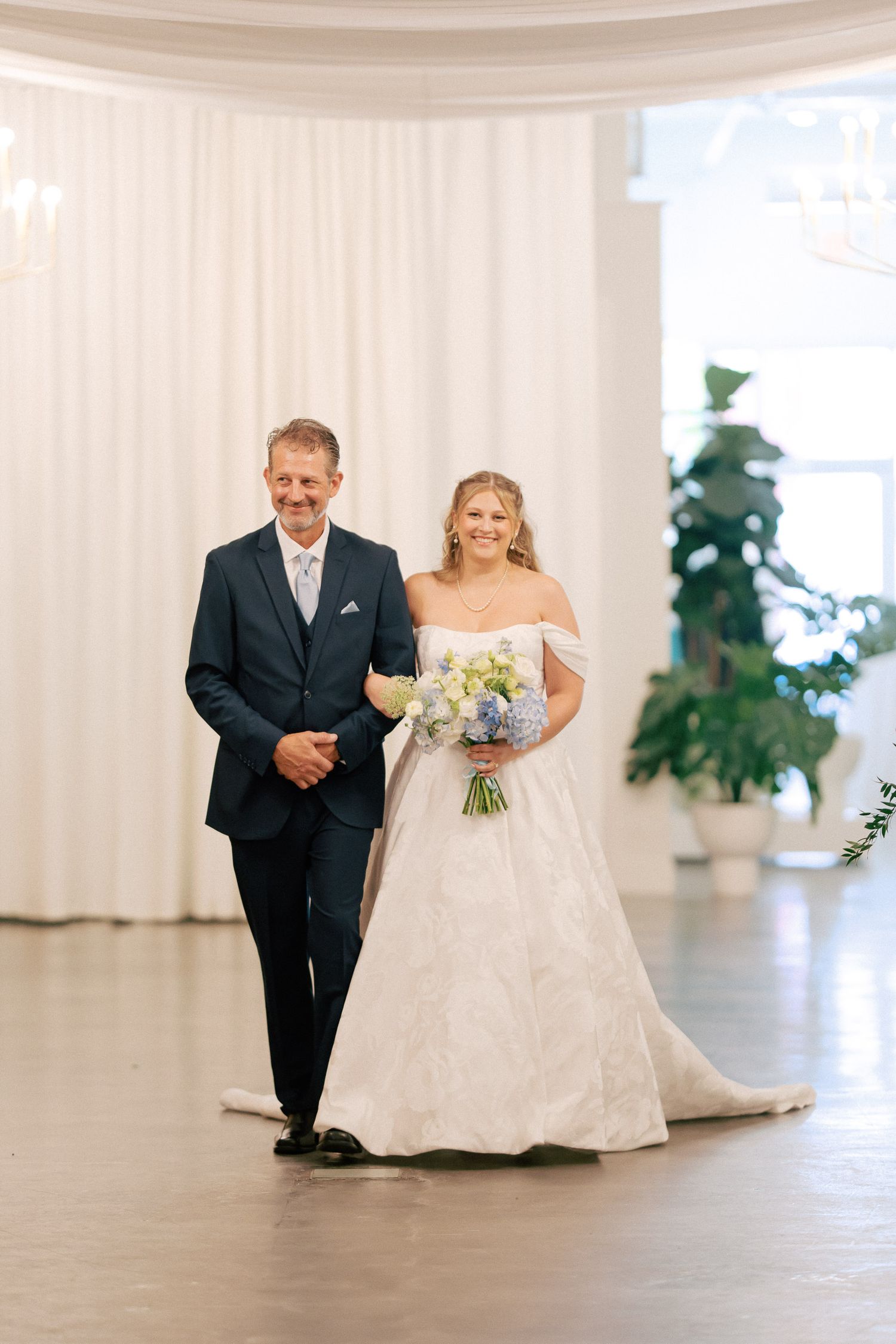Bride in off-shoulder gown walks down aisle with father in navy suit, holding blue and white bouquet.