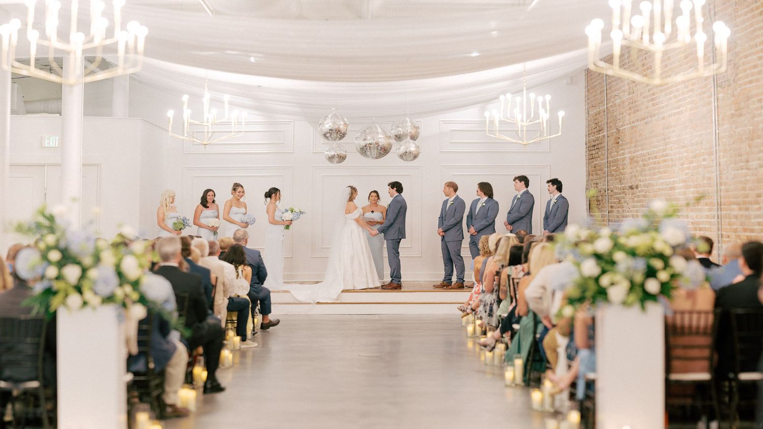 Elegant indoor wedding ceremony with bride and groom at altar, chandeliers, floral arrangements, and seated guests in a white venue.