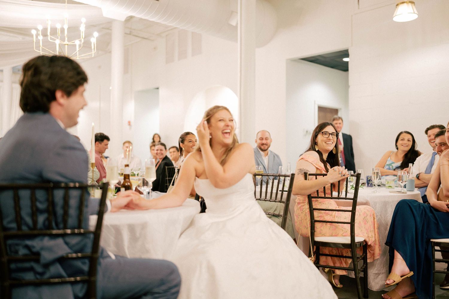 Bride laughing joyfully during wedding reception toast, seated in elegant white venue with guests.