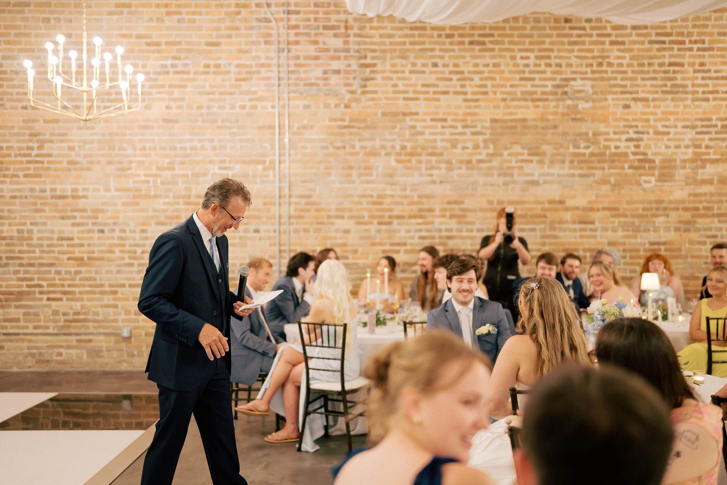 Elegant wedding reception in a brick-walled venue with chandelier, guests seated at tables while a man in navy suit walks.