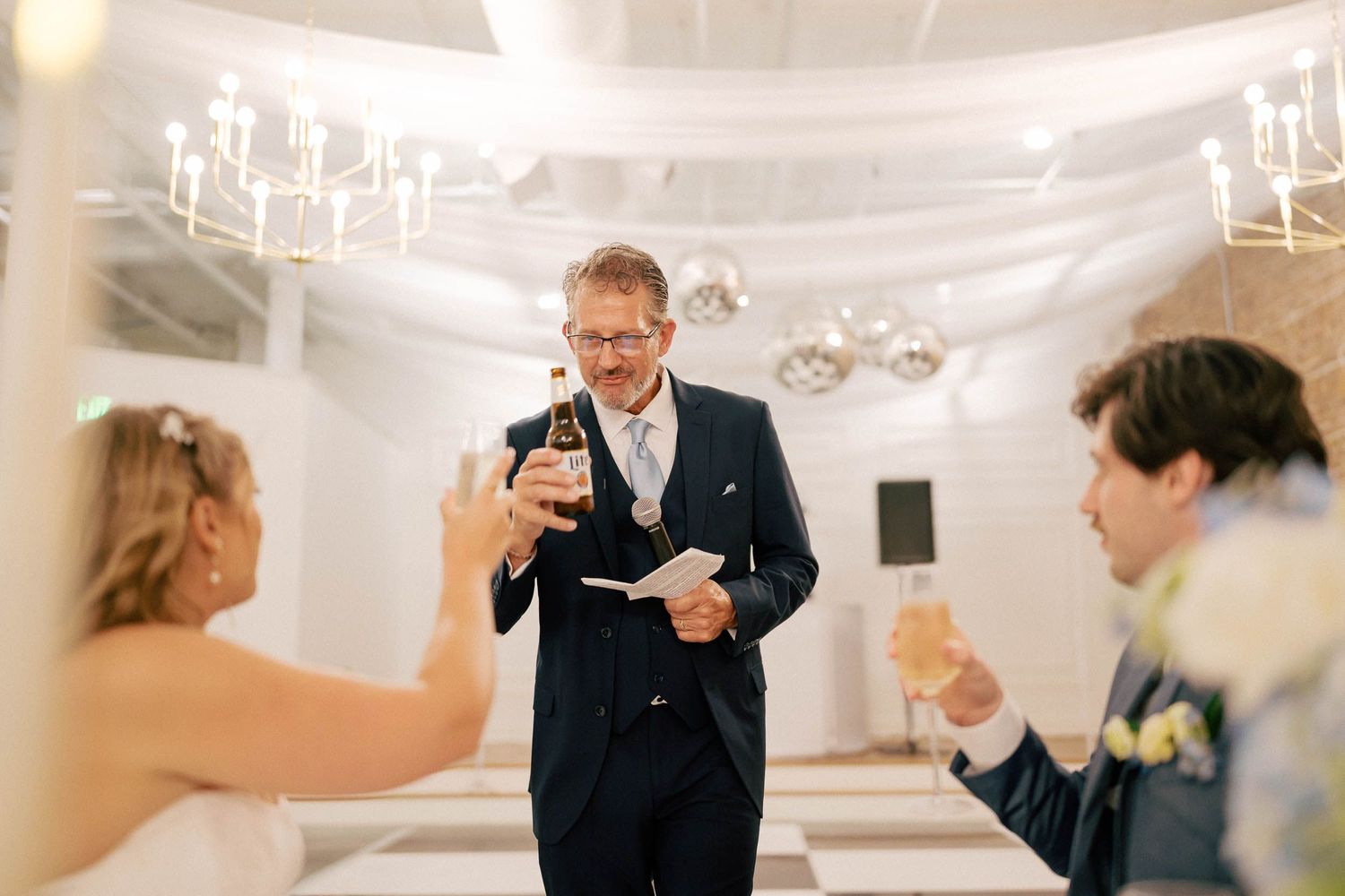 Man in navy suit reads toast at wedding reception while bride and groom raise champagne glasses in elegant venue.