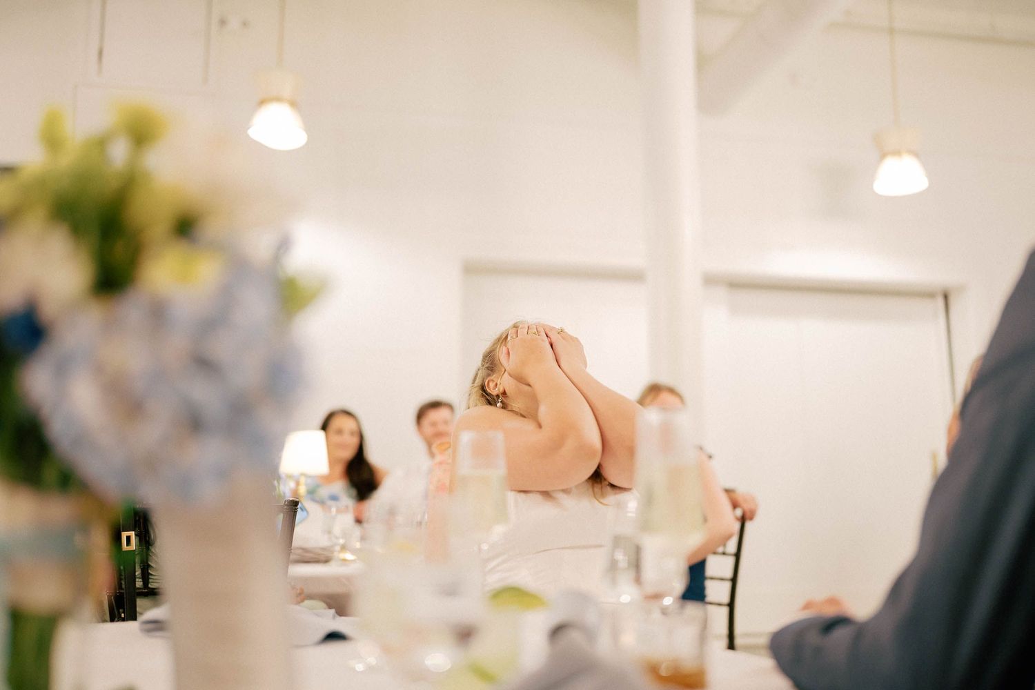 Guest laughing at a wedding reception dinner table in a bright white venue with floral centerpieces.