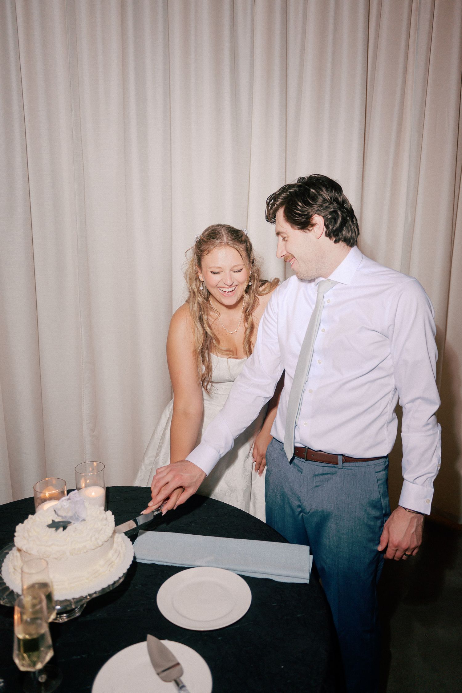 Bride and groom smiling while cutting their white wedding cake together at their elegant reception.