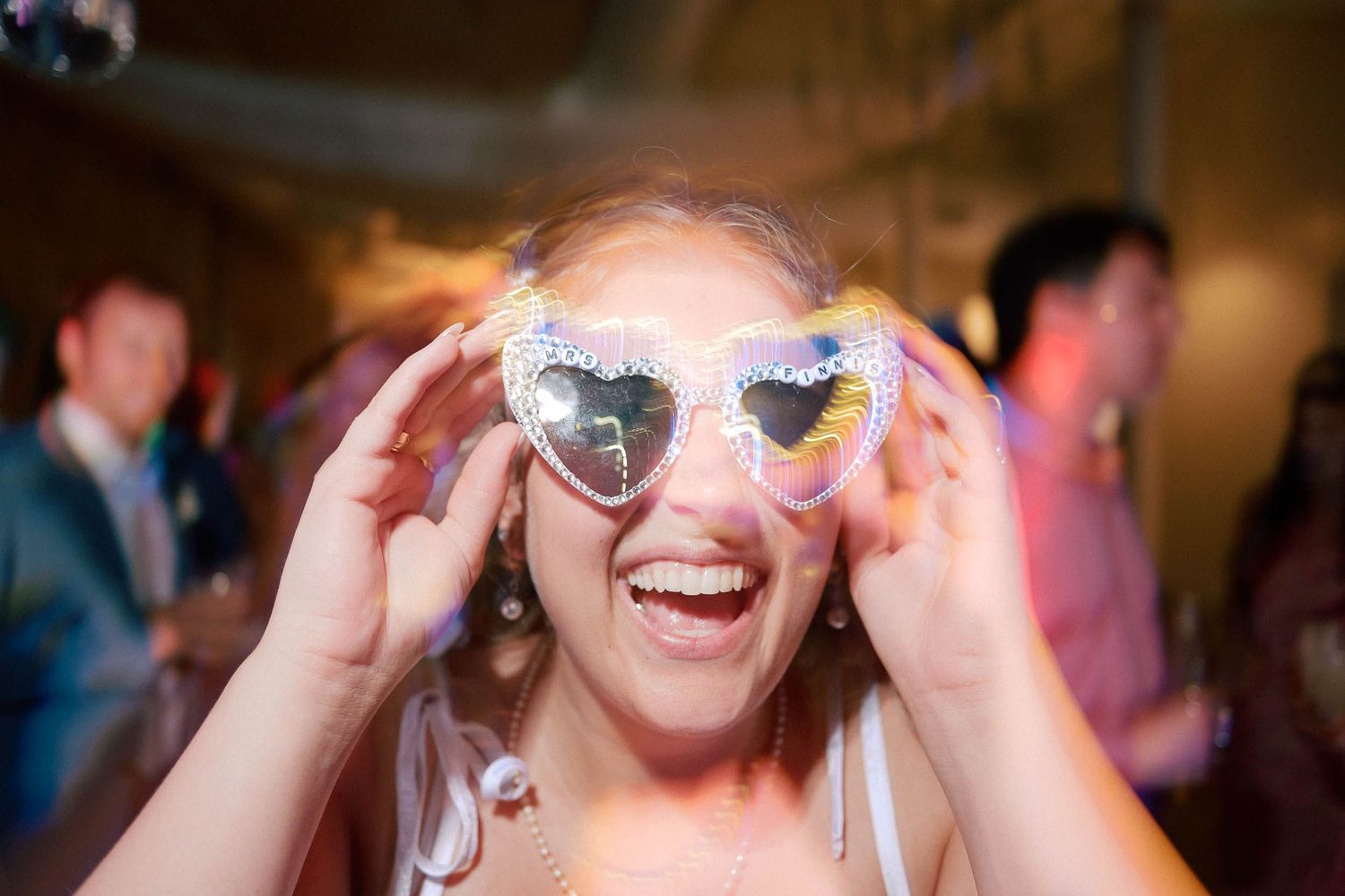 Woman laughing wearing oversized heart-shaped disco mirror sunglasses at a wedding reception party.
