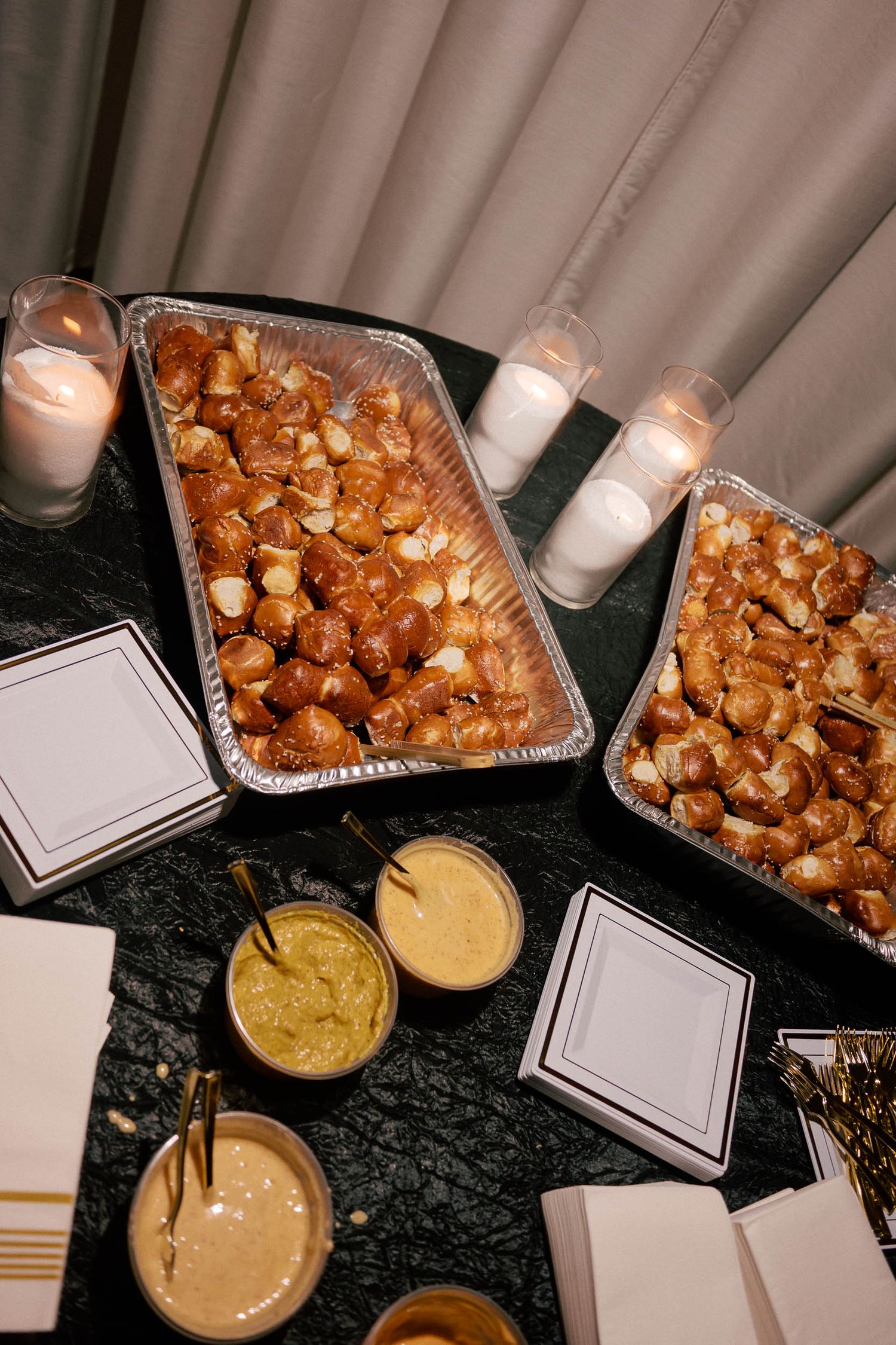 Two trays of golden pretzel bites served with assorted mustard dipping sauces on a black sequin table.