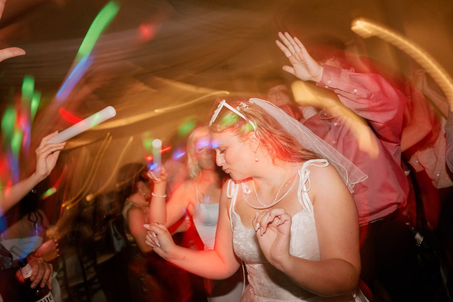Bride dancing joyfully at wedding reception surrounded by guests holding colorful glow sticks.