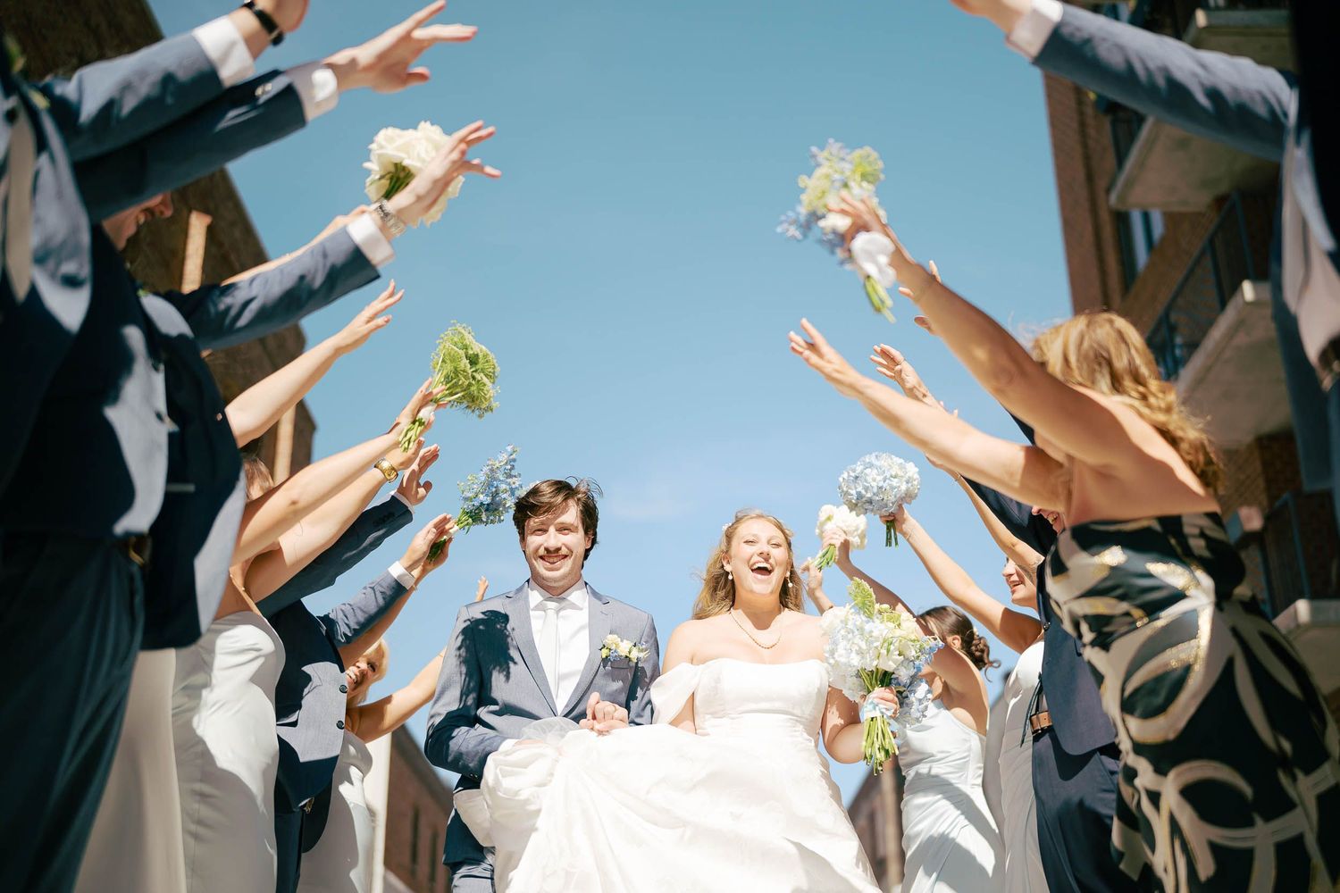 Joyful newlyweds walk under raised arms of wedding party outdoors on a sunny day.