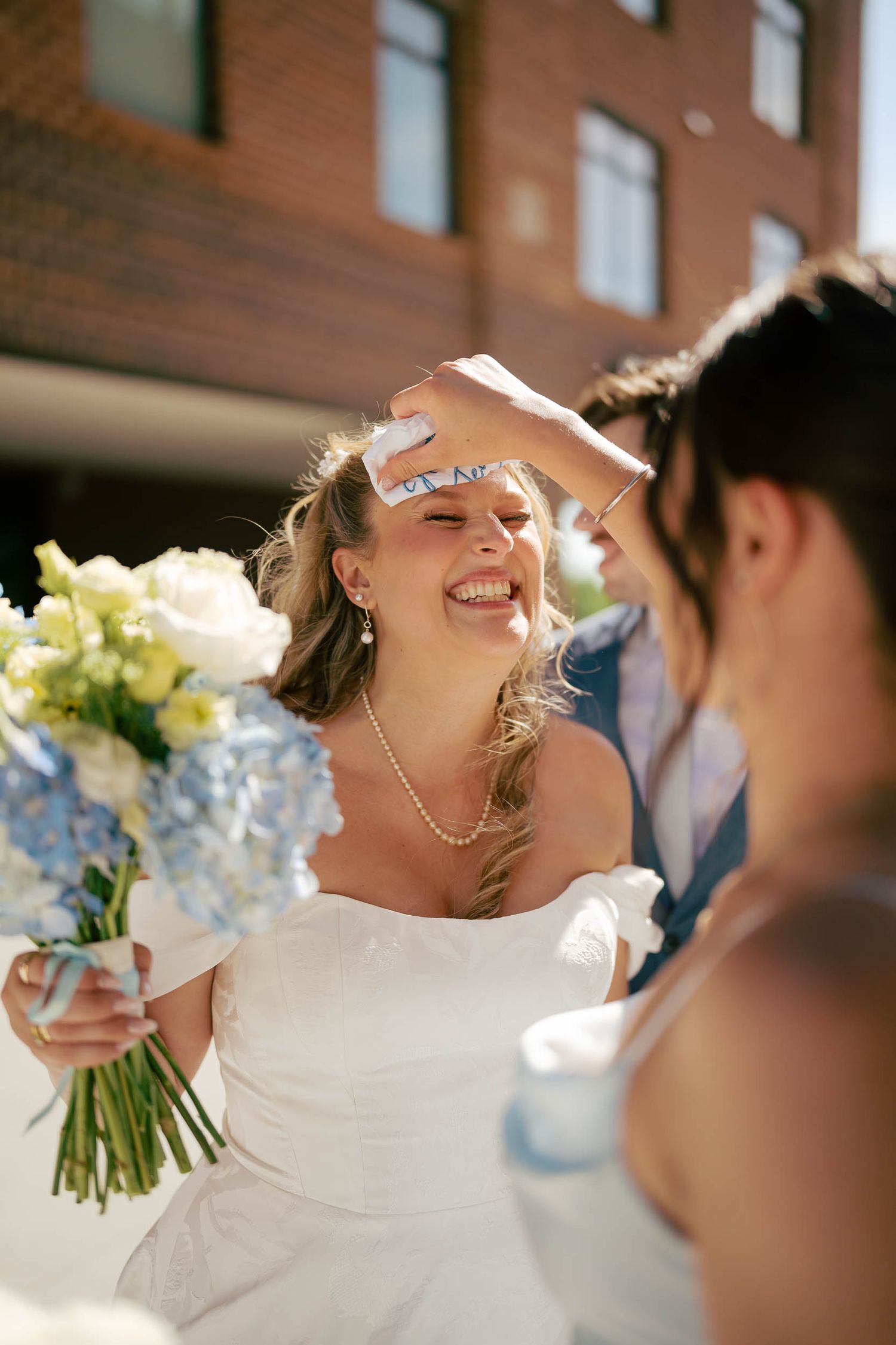Smiling bride in off-shoulder gown holds blue and white bouquet outdoors on sunny wedding day.