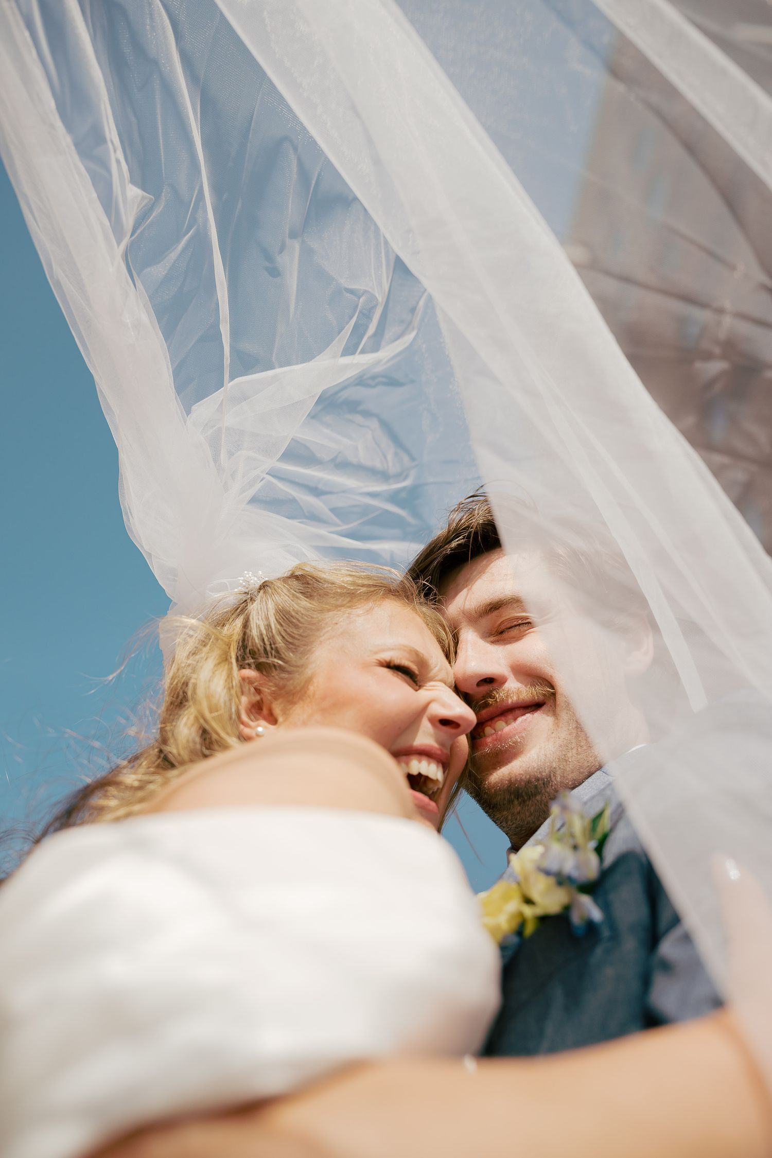 Joyful bride and groom laughing together under flowing white veil against blue sky on their wedding day.