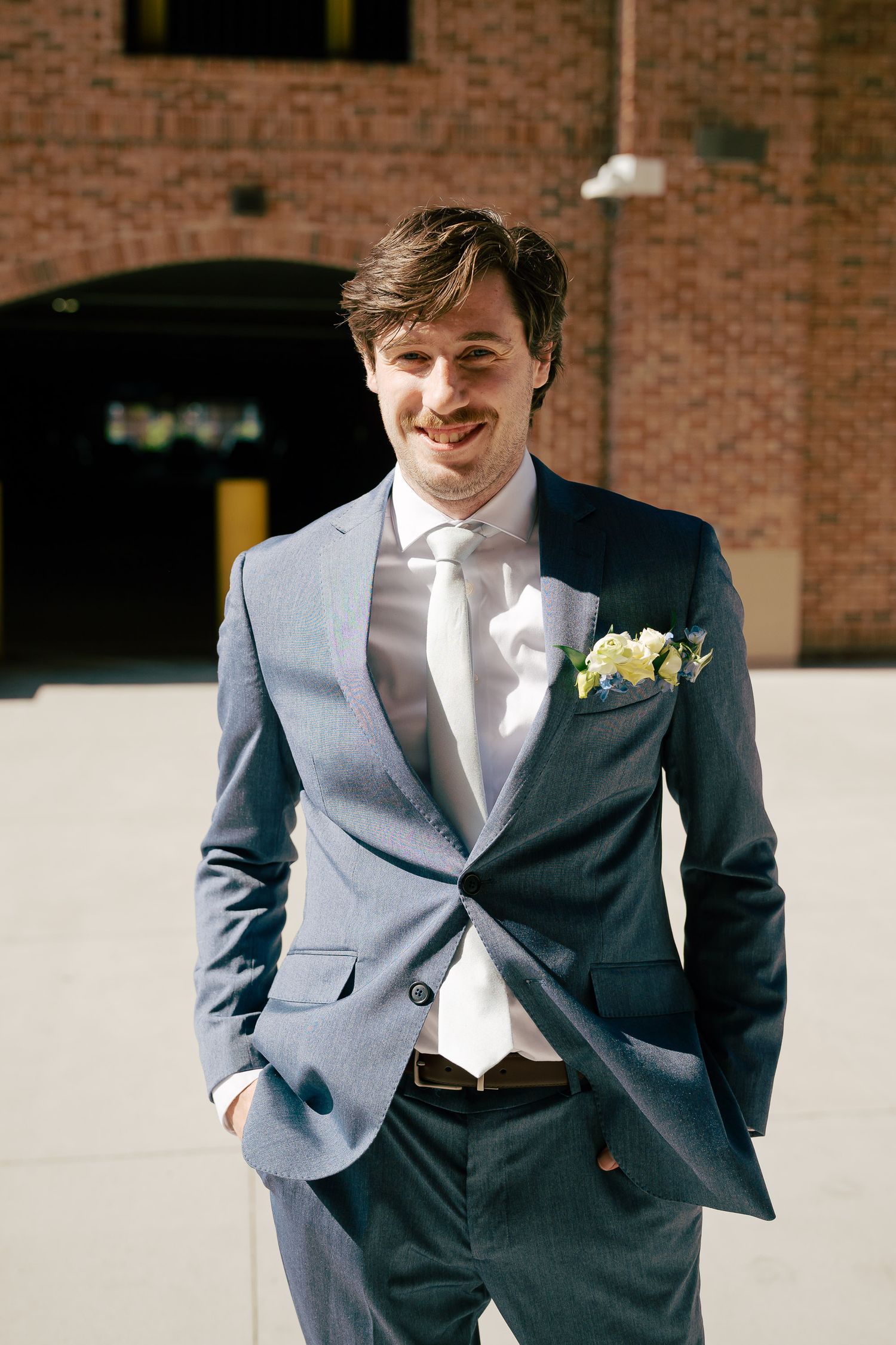 Smiling groom in navy blue suit with white floral pocket square posing outdoors on sunny wedding day.