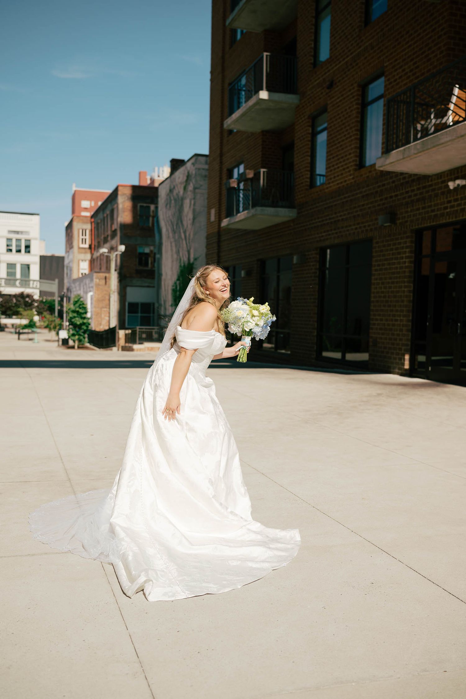 Bride in off-shoulder white gown with train and bouquet posing outdoors on sunny day near brick building.