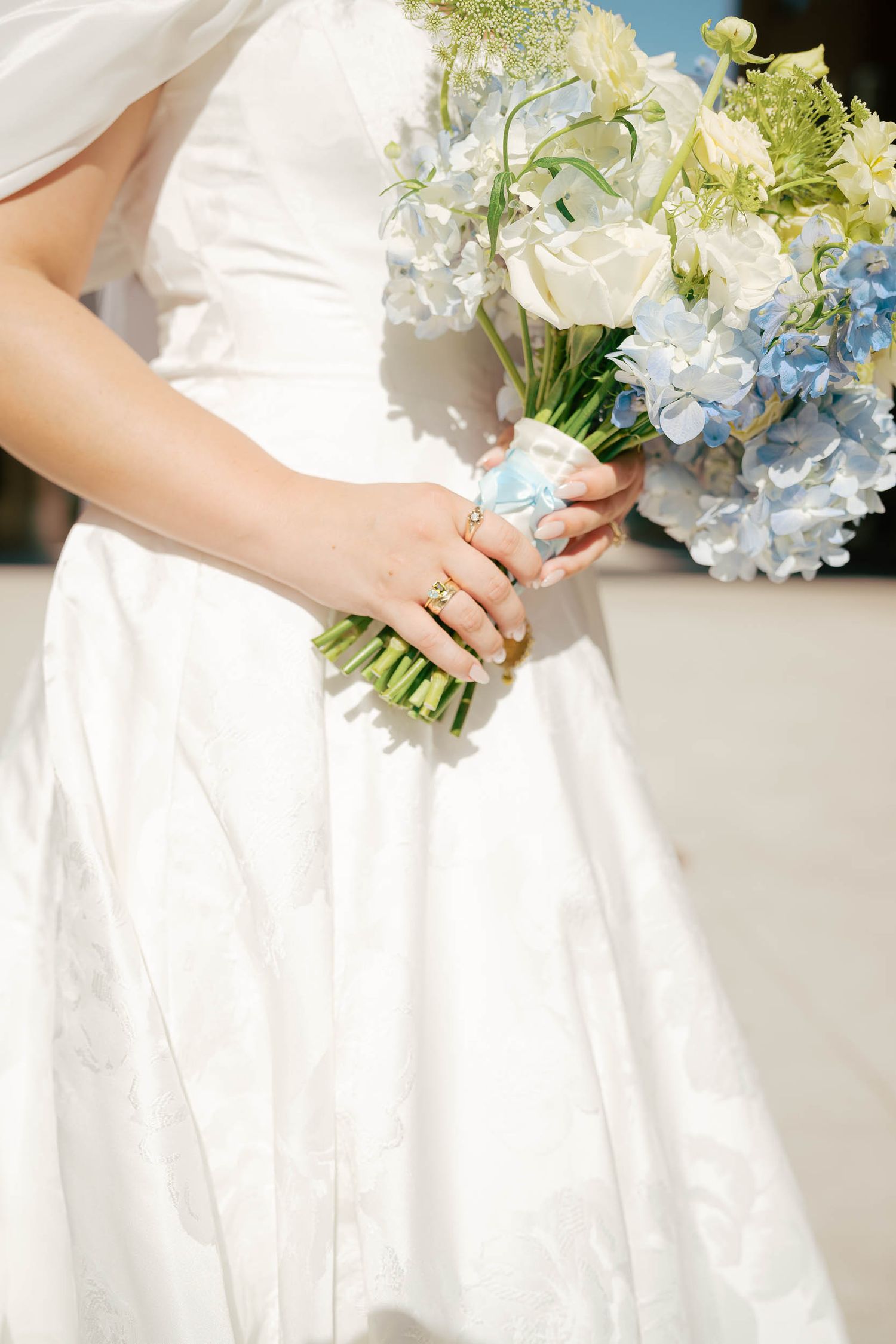Bride in white gown holding blue hydrangea and white rose bouquet tied with light blue ribbon.